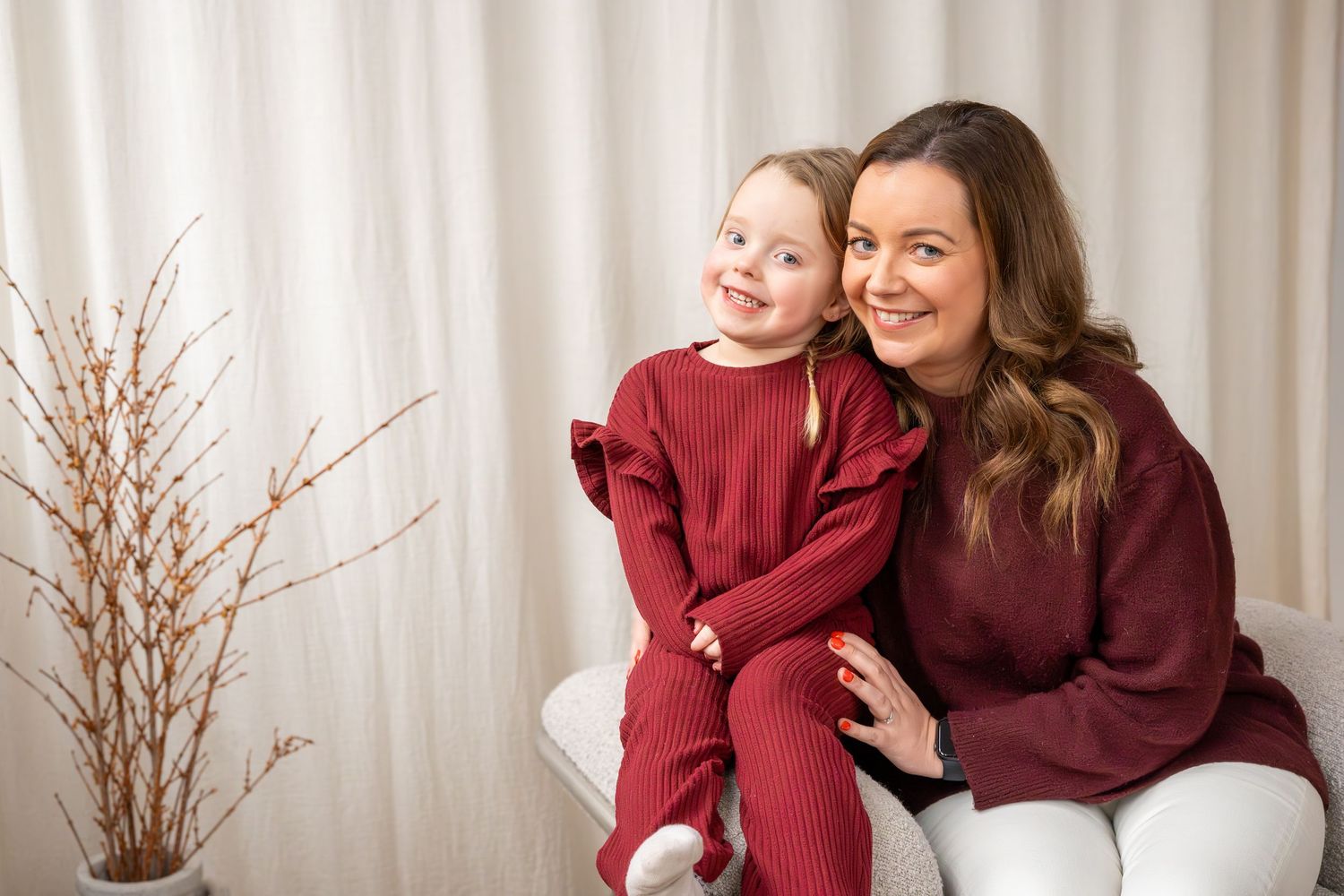 A mother and young daughter wearing matching burgundy outfits pose together on a white chair.