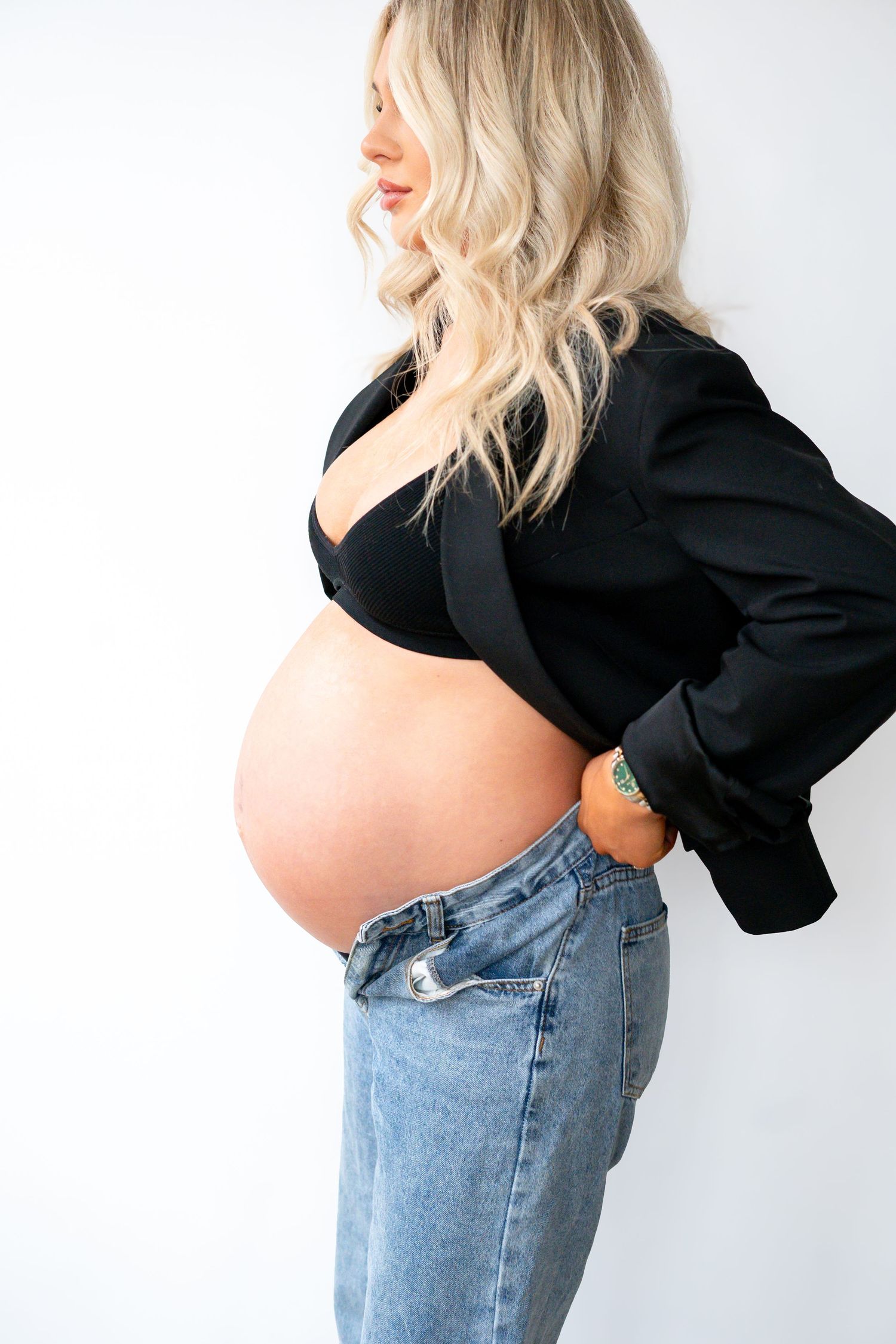 Pregnant blonde woman in black blazer and jeans showing baby bump, side profile view.