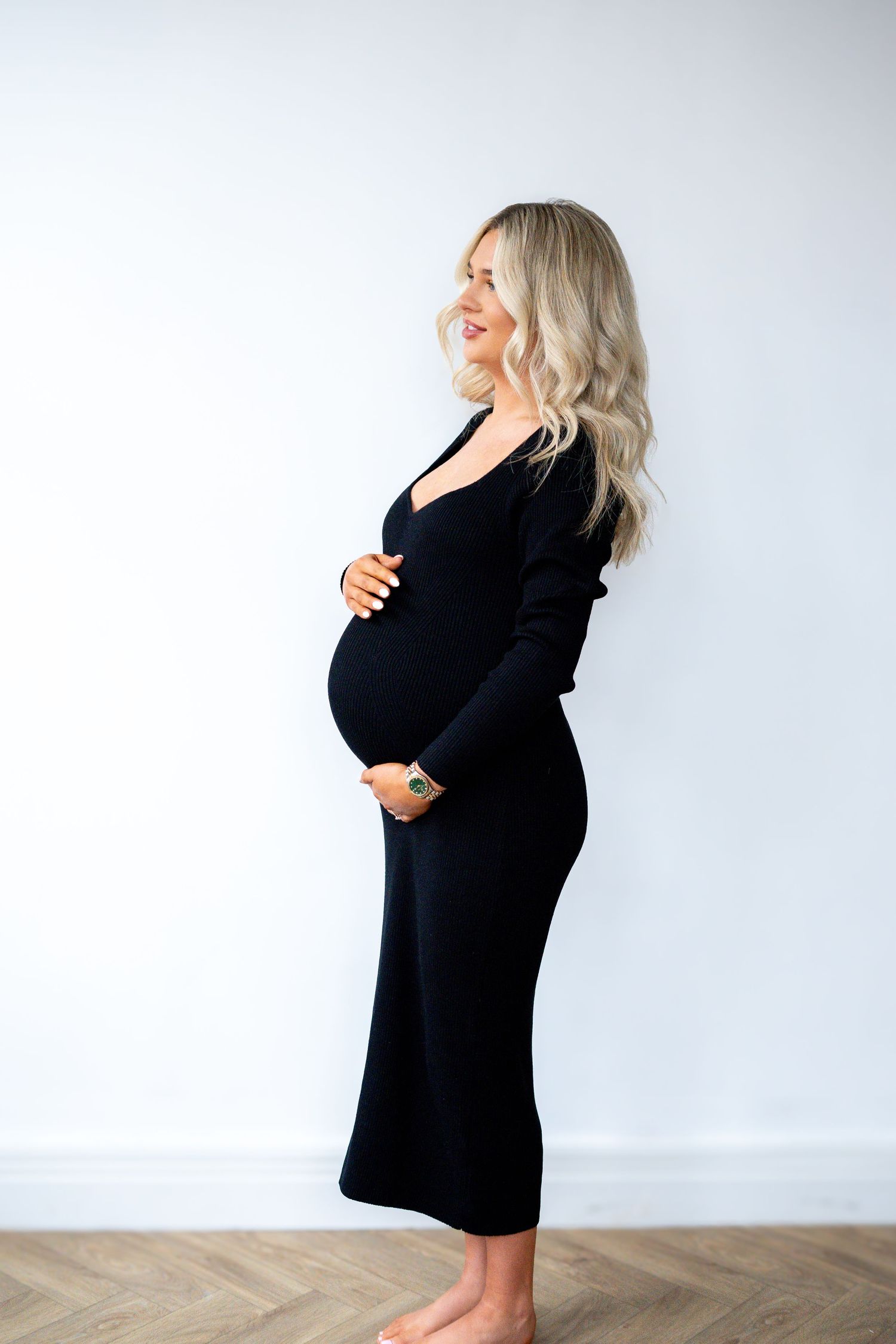 Joyful pregnant woman in black ribbed midi dress smiling sideways, barefoot on herringbone floor.