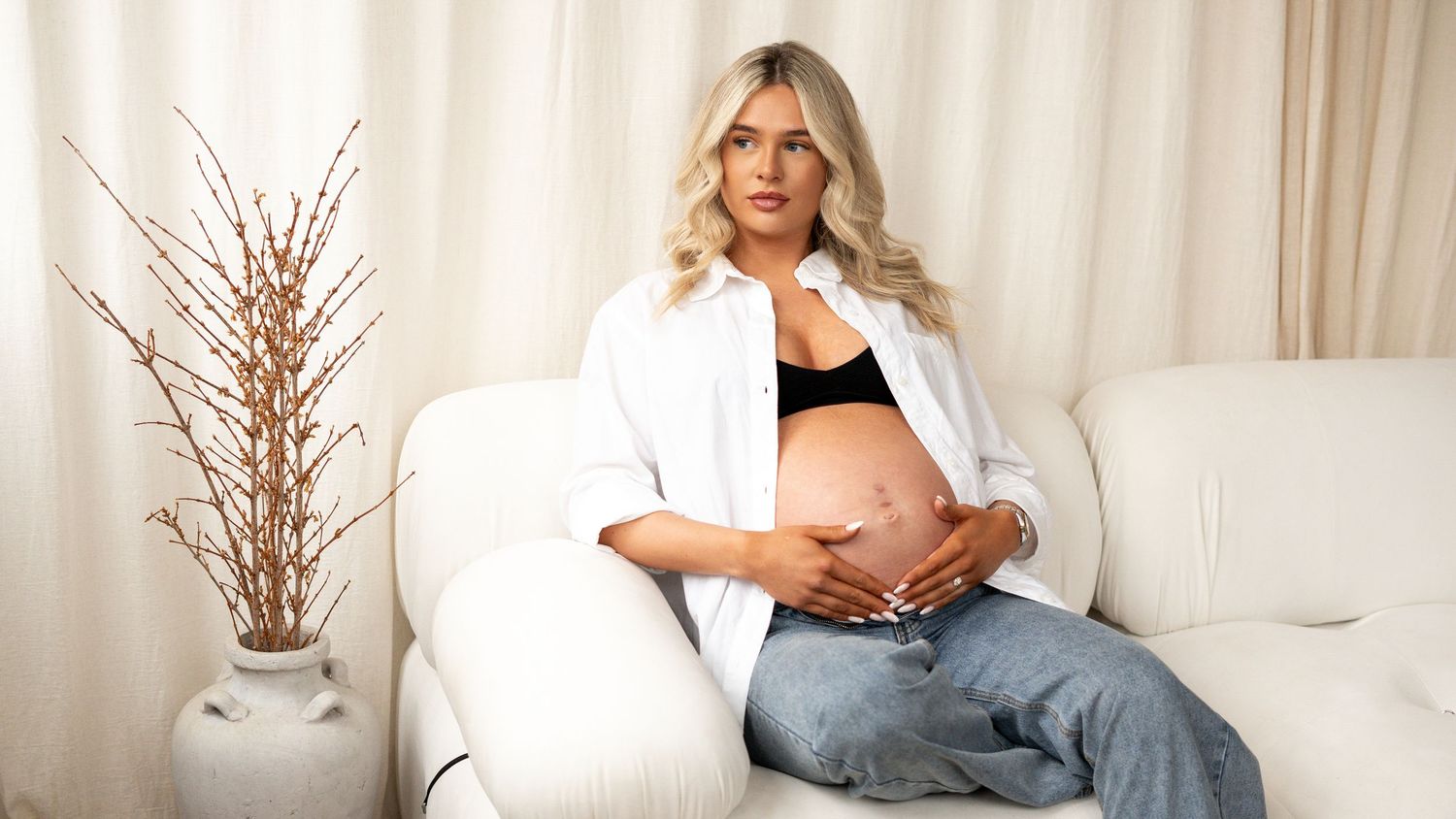 Blonde pregnant woman in white shirt and jeans sitting on white sofa holding baby bump in bright room.