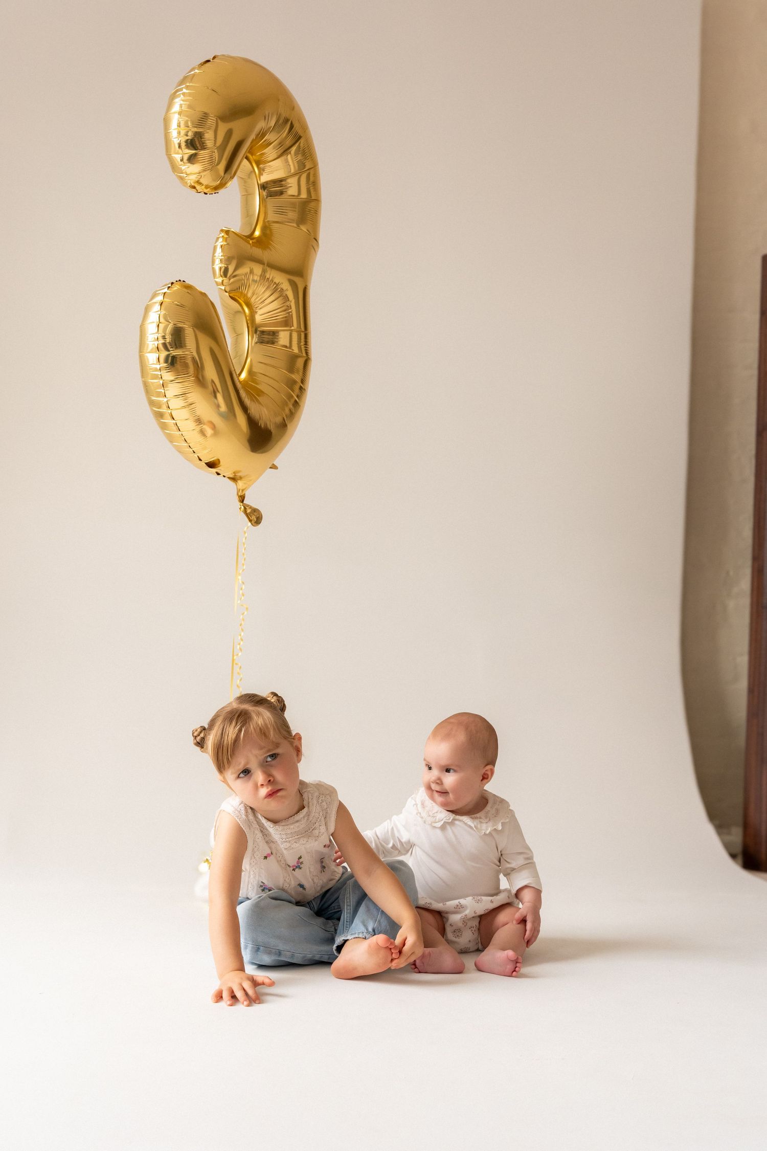 Two young children sit on white backdrop with gold number 3 balloons, celebrating a third birthday.