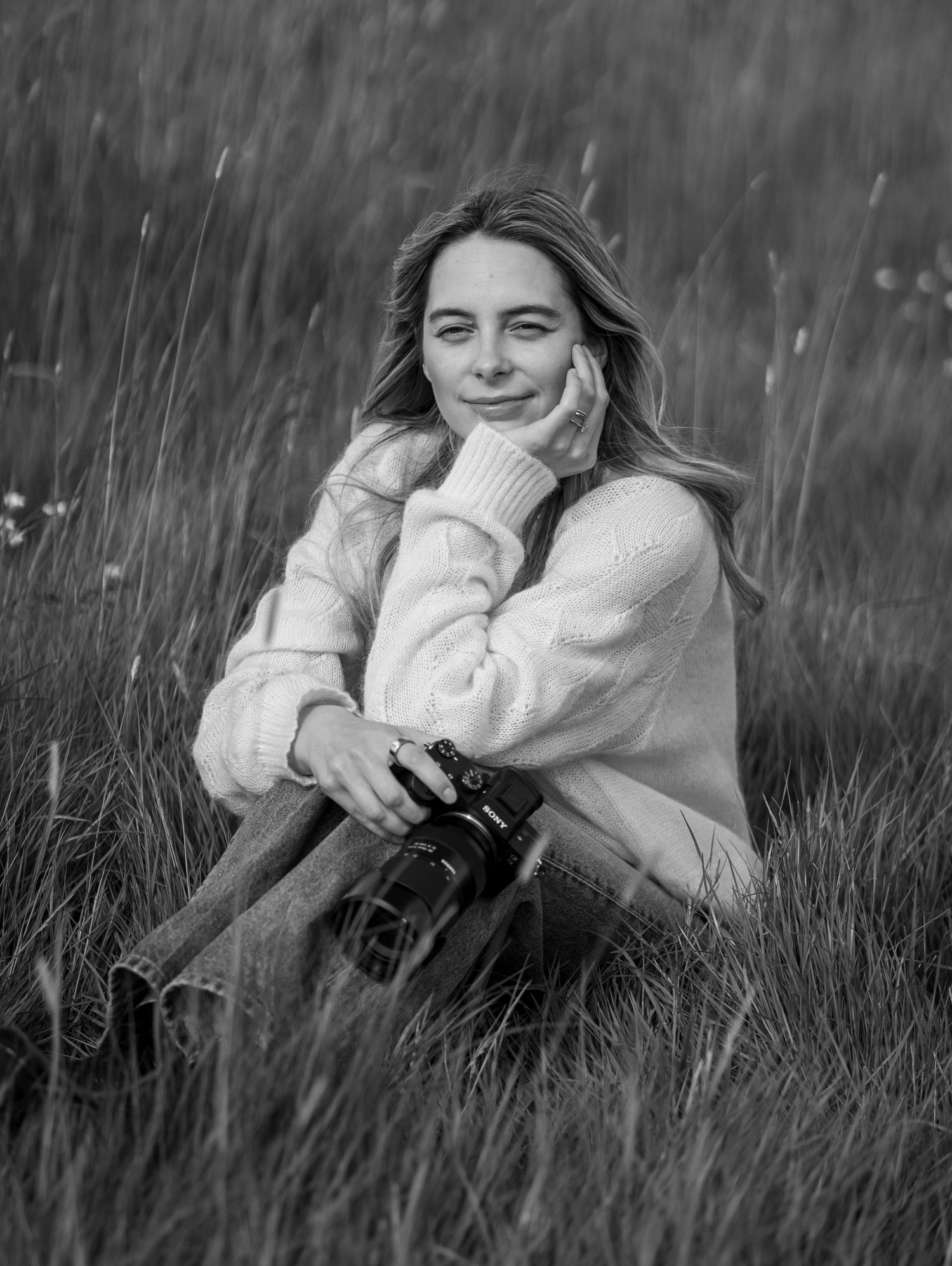 Young woman in cream sweater sitting in tall grass field, smiling and holding a camera in black and white photo.