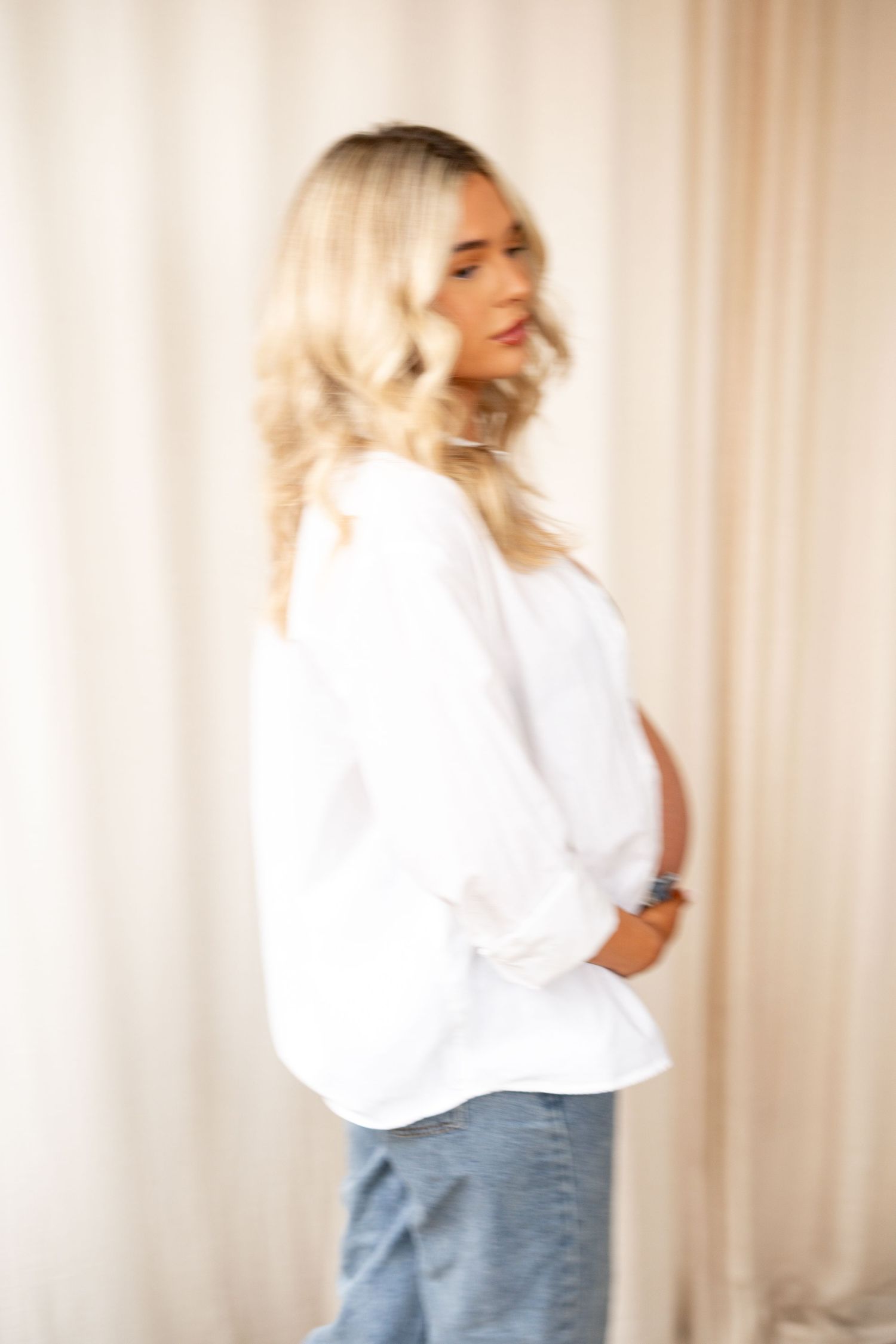 Side profile of pregnant blonde woman in white shirt and jeans with soft curtain backdrop.