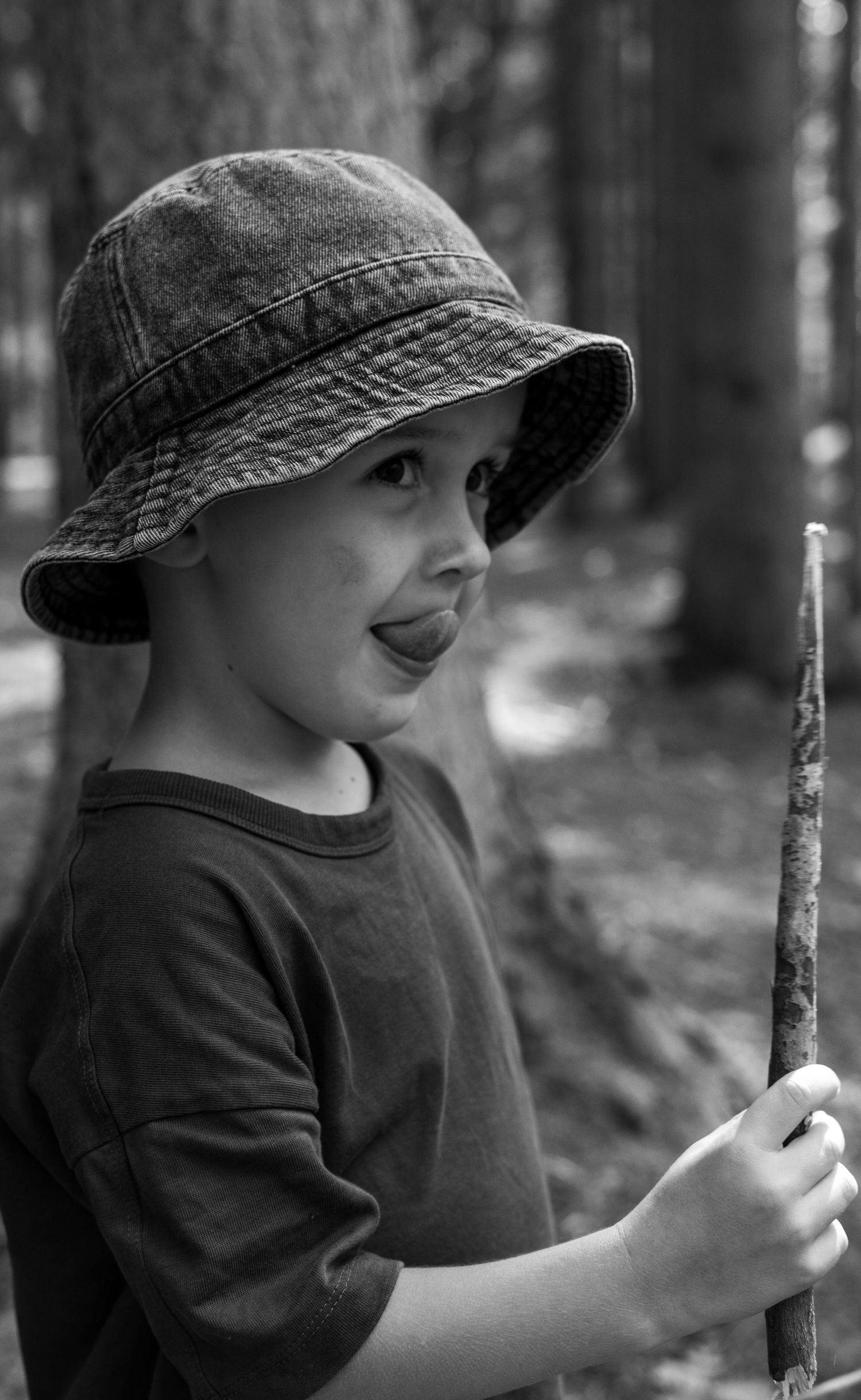 Black and white photo of young boy wearing bucket hat holding a stick in forest setting.