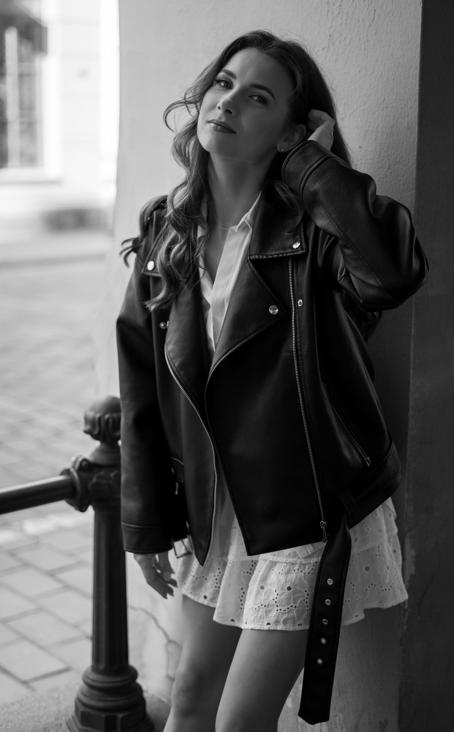 Woman in black leather biker jacket and white mini dress leaning against wall in black and white photo.