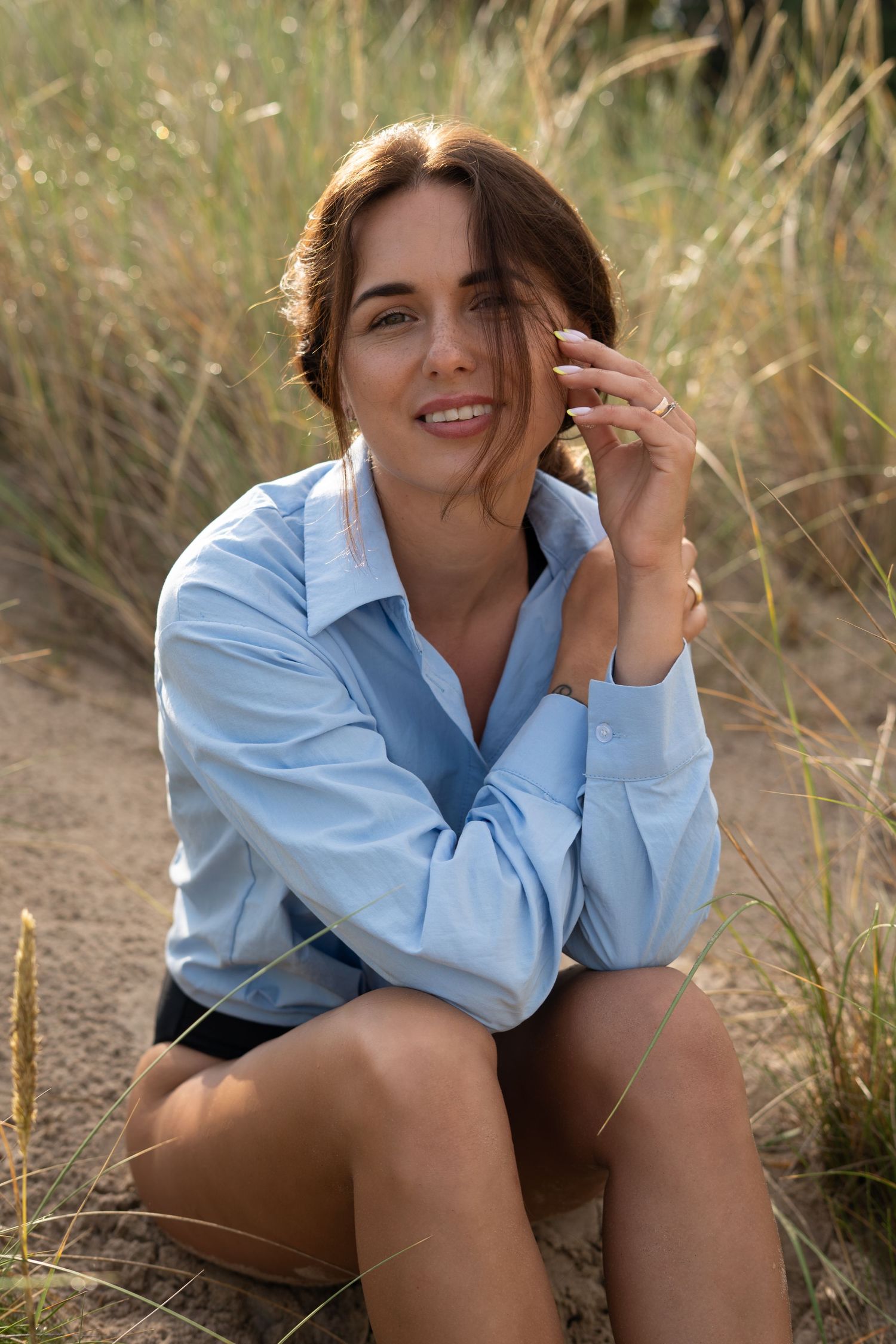 Young woman in blue shirt sitting on sandy dunes smiling at camera with tall grass in background.