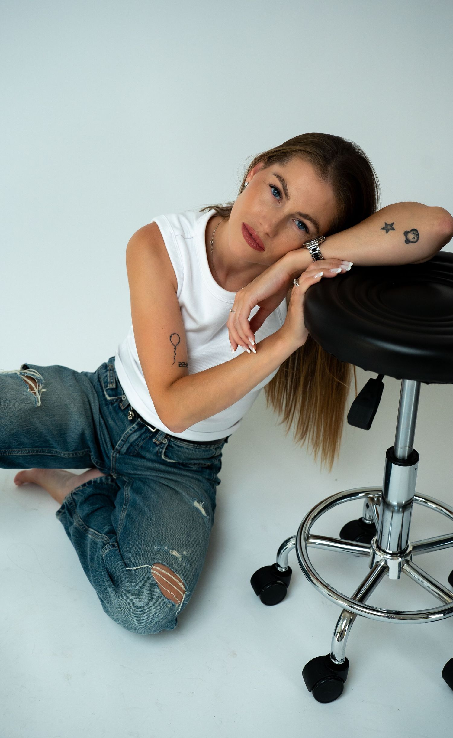 Tattooed woman in white top and ripped jeans sits on floor leaning against black stool on white background.