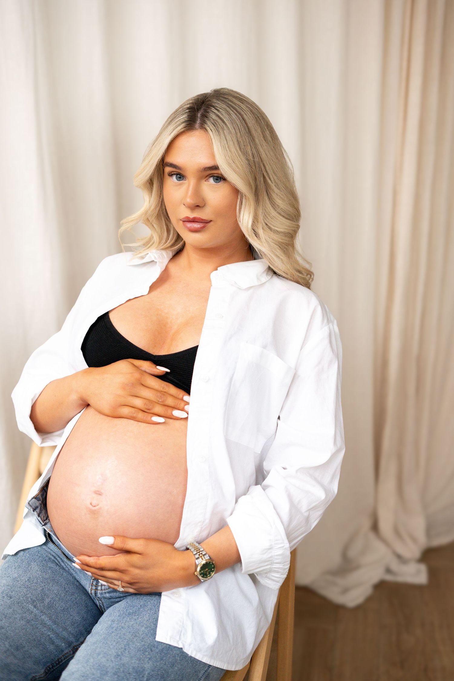 Pregnant blonde woman in white shirt and jeans sitting on chair holding baby bump.