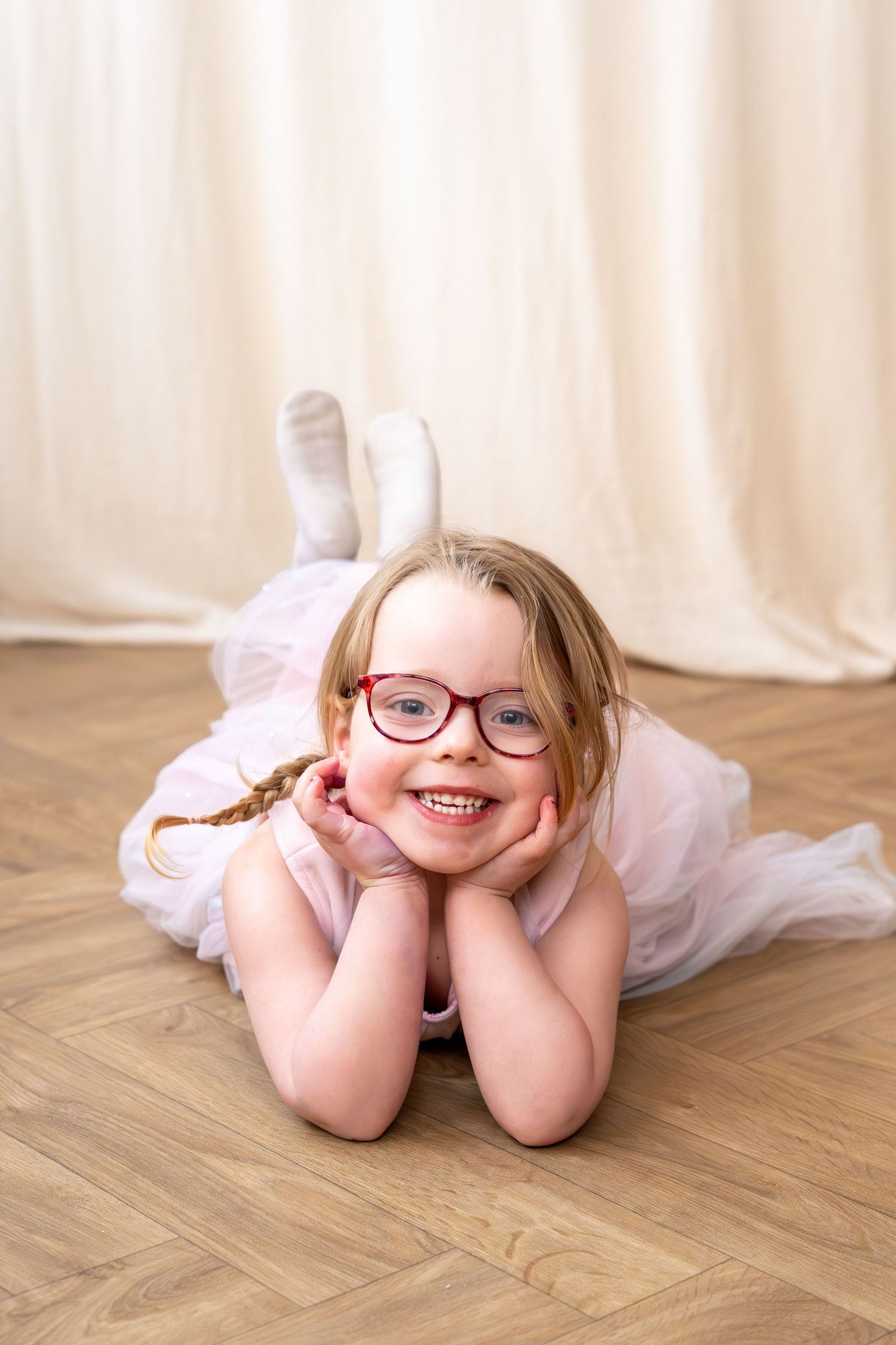 Cheerful young girl in pink tutu and red glasses lying on wooden floor smiling at camera.