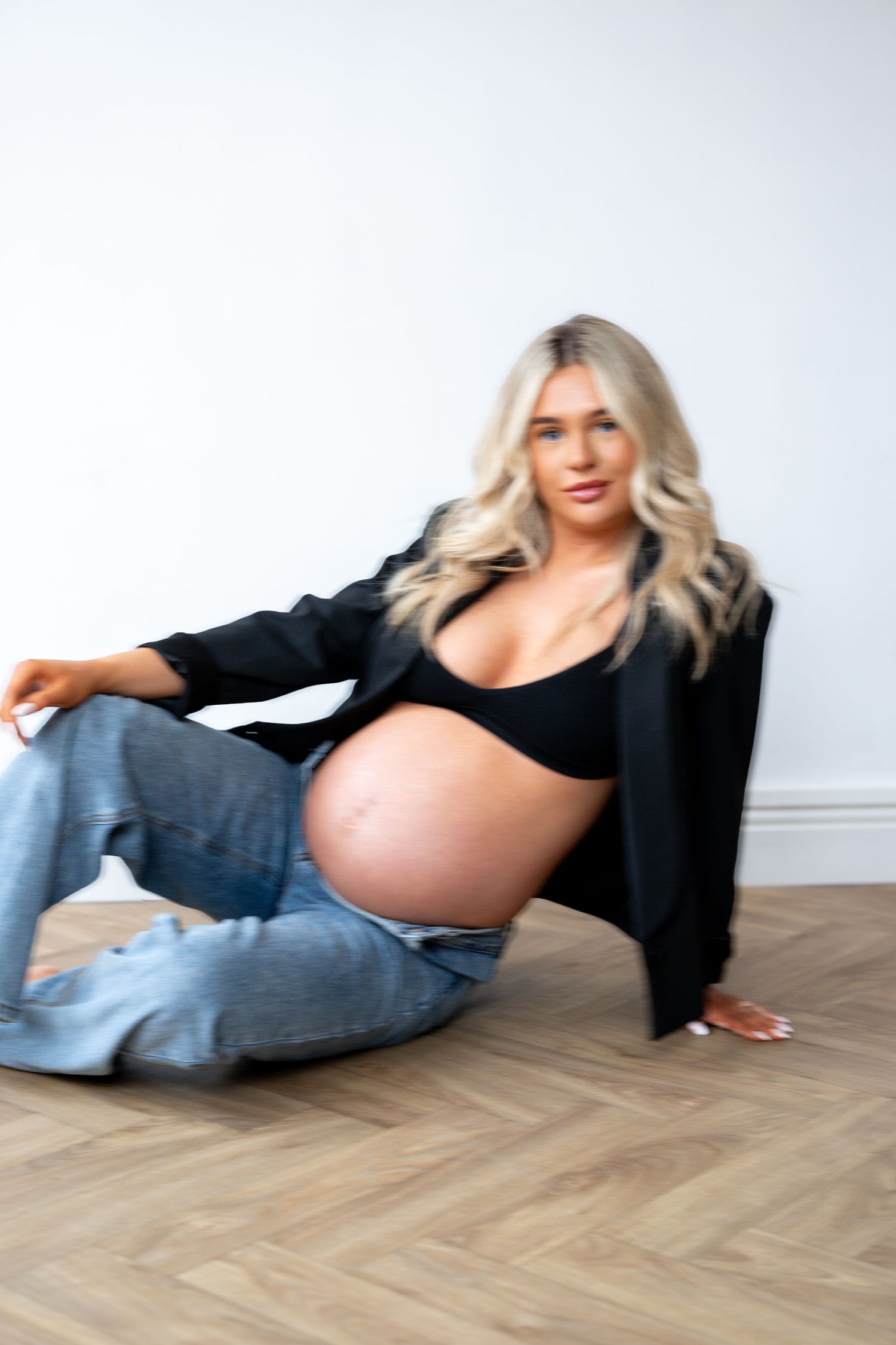 Pregnant blonde woman sitting on floor showing bare bump, wearing black blazer and blue jeans.