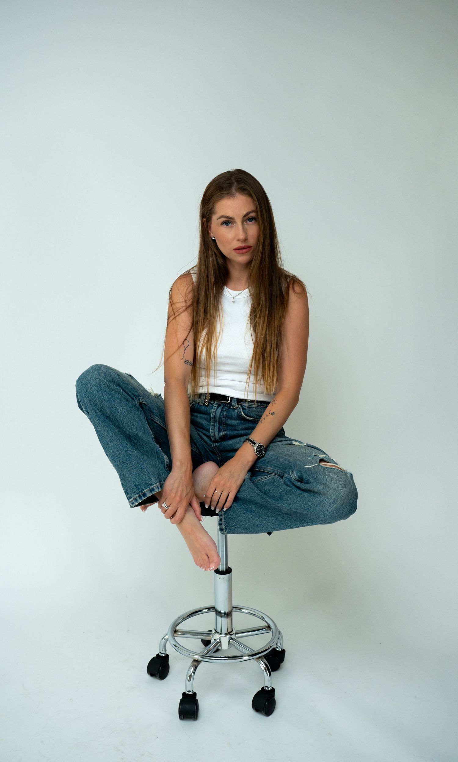 Young woman in white crop top and wide-leg ripped jeans sitting barefoot on chrome stool.