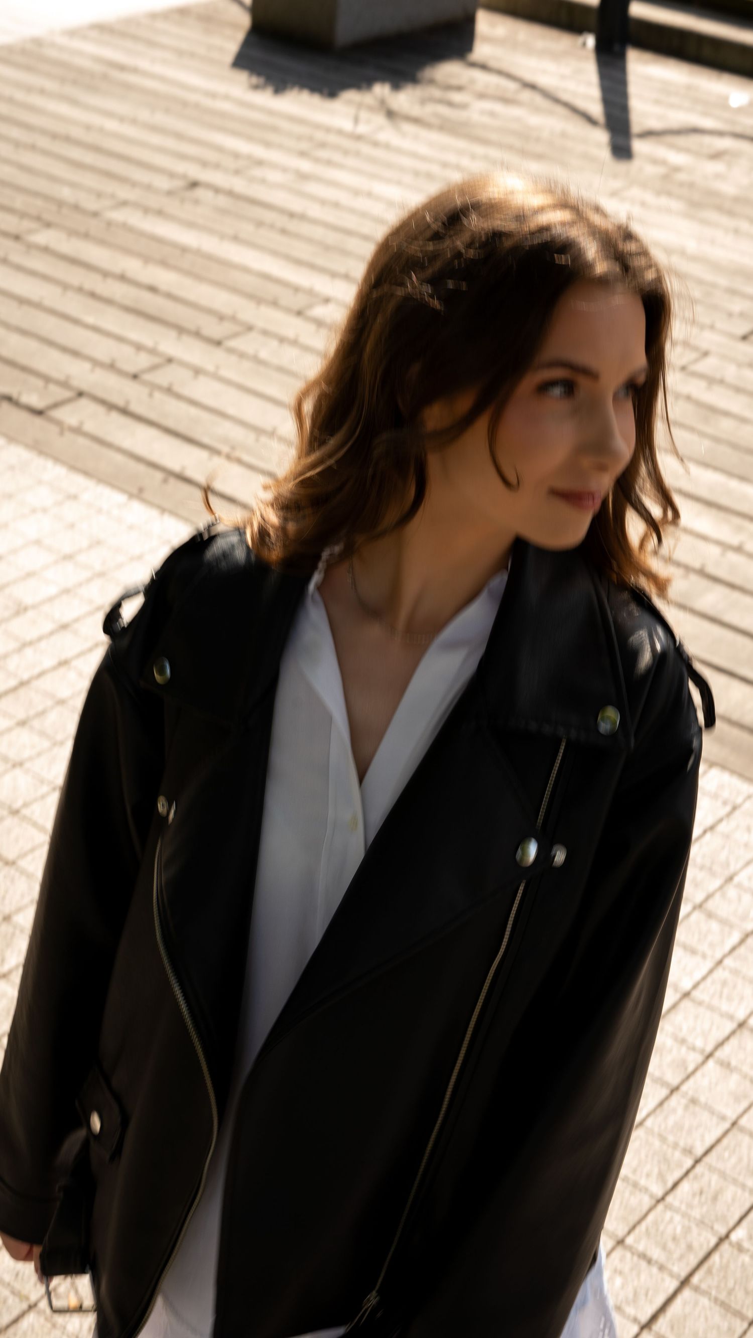 Young woman in black leather biker jacket over white dress stands in sunlight on wooden pavement.