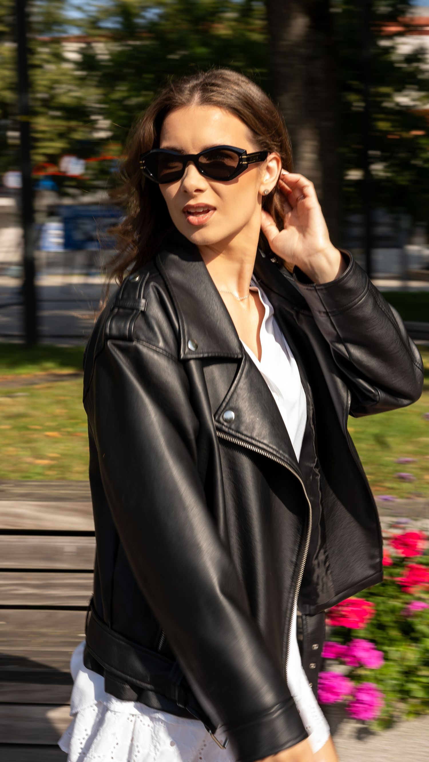 Woman in black leather jacket and black sunglasses adjusting hair outdoors near colorful flowers in park.