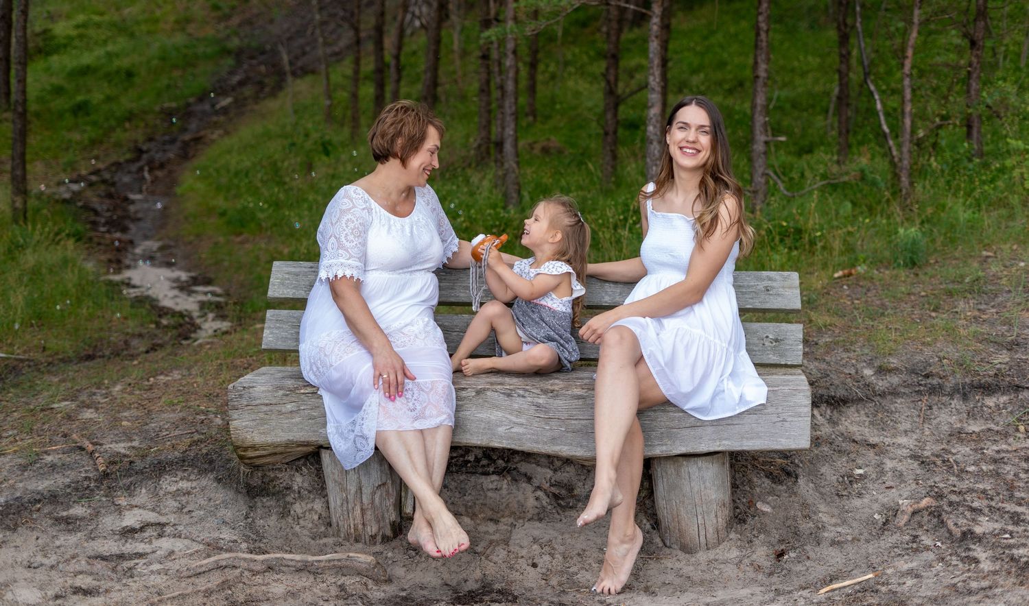 Two women and a child sit on a wooden bench outdoors, sharing a joyful moment together.