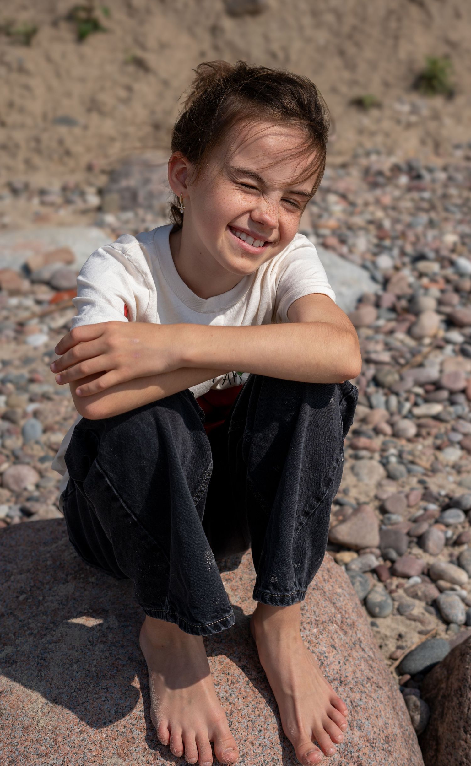 A barefoot child smiling and sitting cross-legged on rocks at a pebble beach in a white t-shirt.