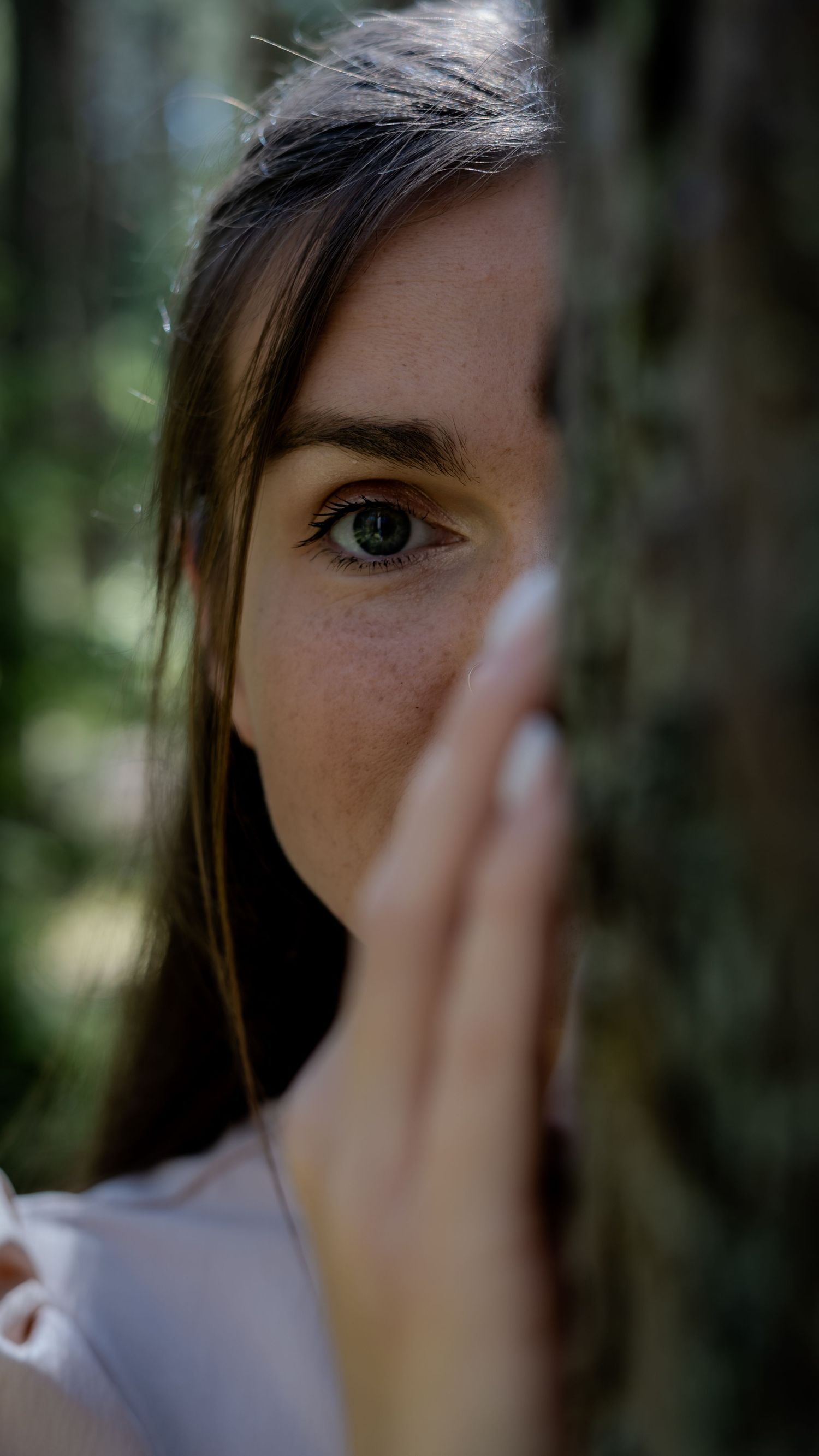 Close-up portrait of young woman with green eyes and freckles partially hidden behind a tree trunk outdoors.