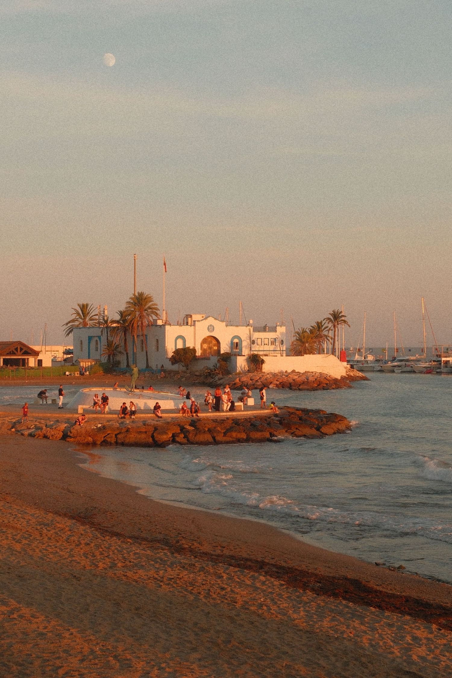Sunset view of a white Mediterranean building on a rocky coastal promontory with palm trees, sandy beach, and a full moon rising.