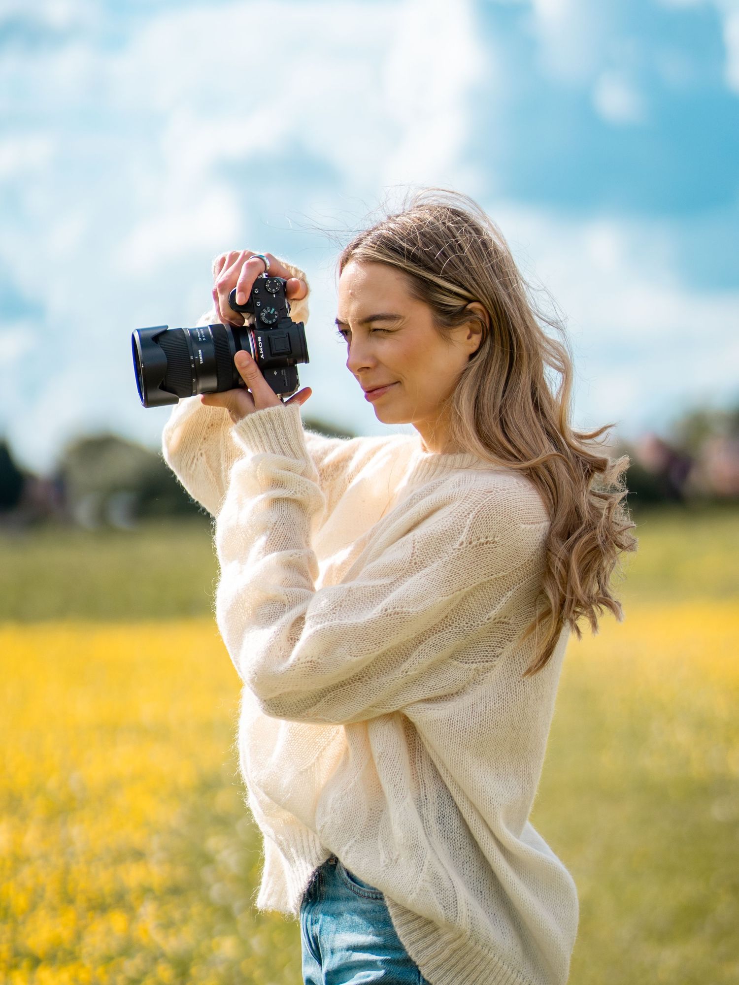 Blonde woman in cream sweater photographs with DSLR camera in yellow flower field.