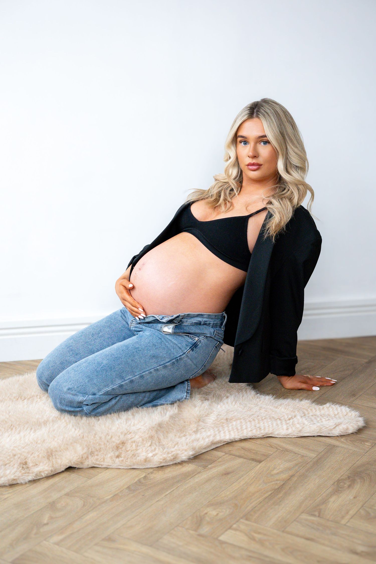 Pregnant woman kneeling on fur rug in black blazer and jeans, cradling bare baby bump.