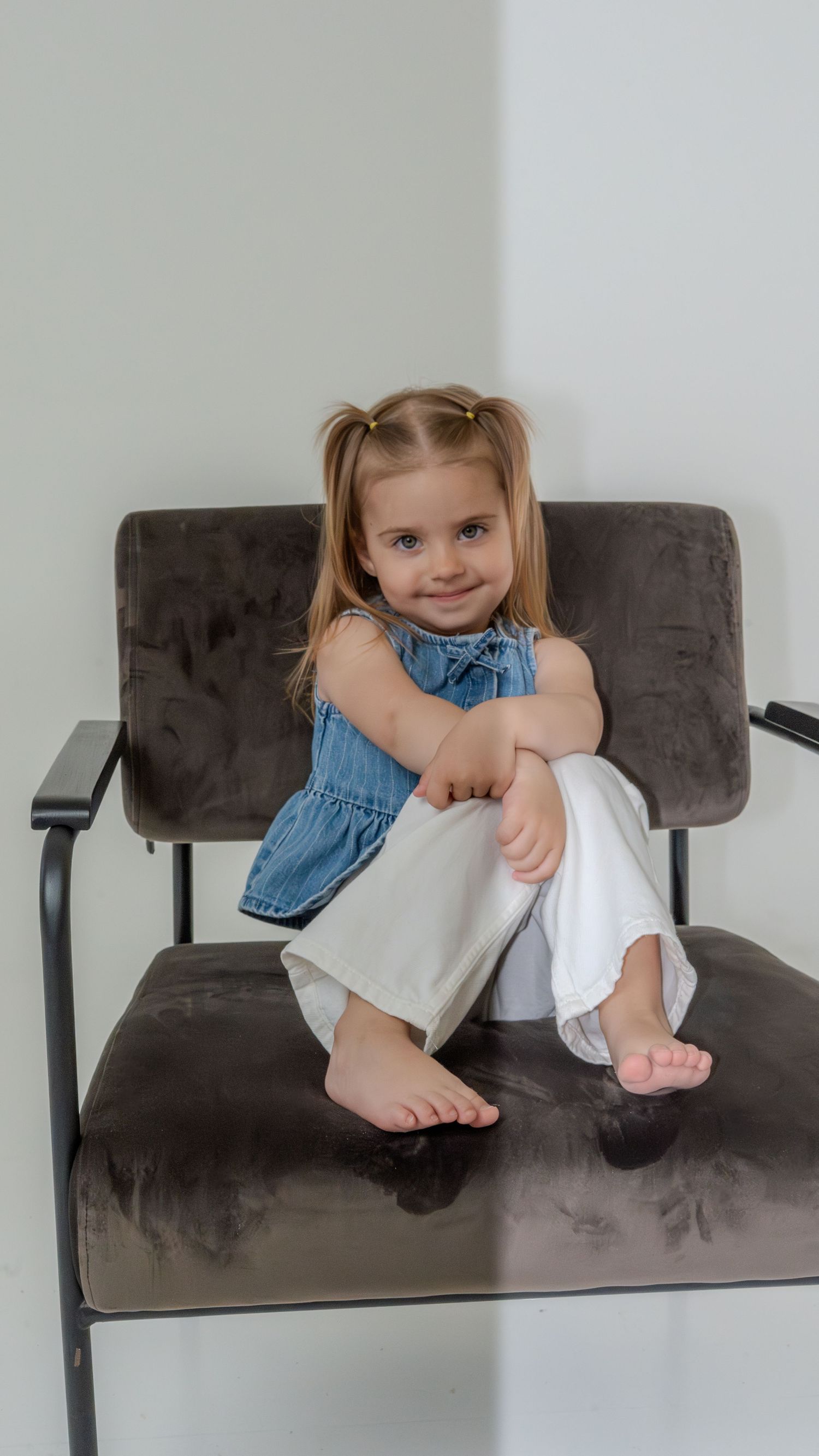 A cute blonde toddler girl in a blue top and white pants sits curled up in a gray velvet chair.