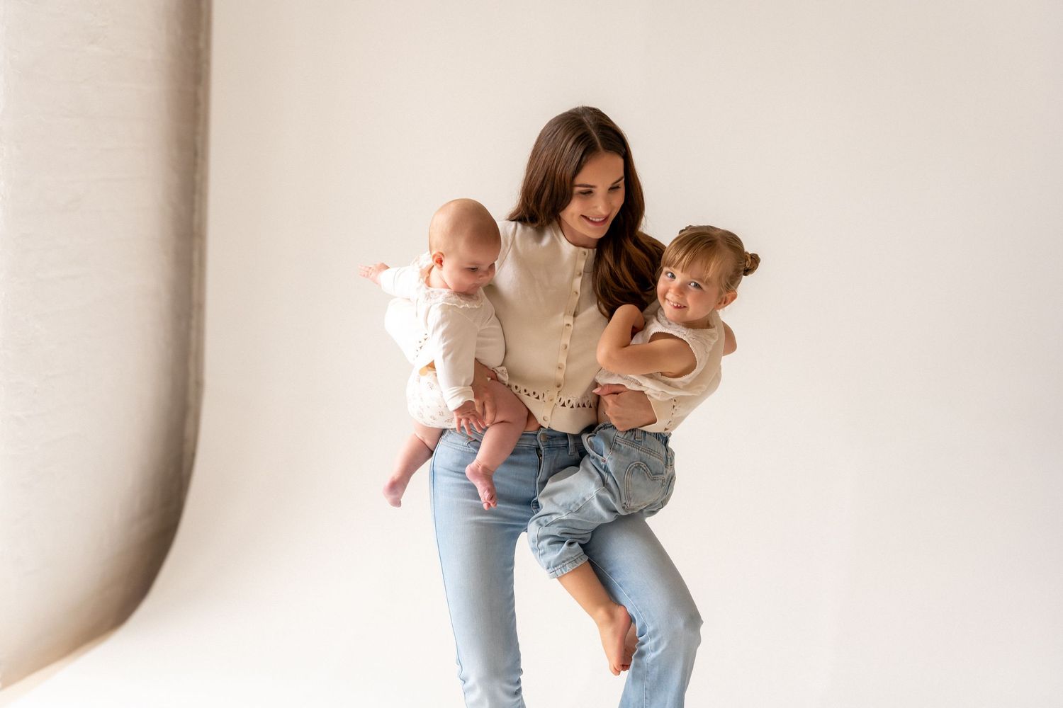 A smiling brunette woman in jeans holds two young toddlers against a white studio backdrop.