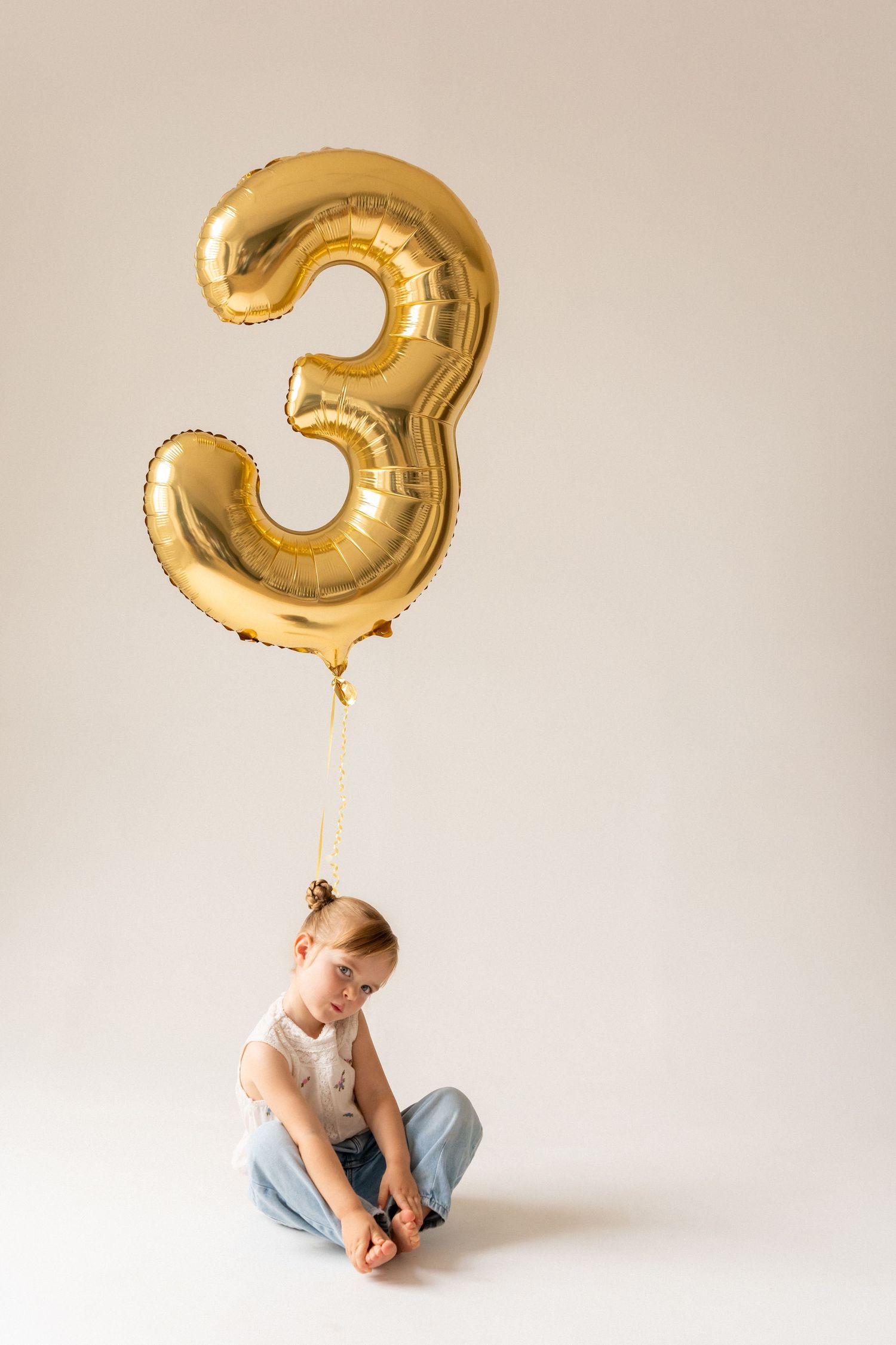 A young girl sits on the floor beneath a large gold number 3 balloon, celebrating her third birthday.