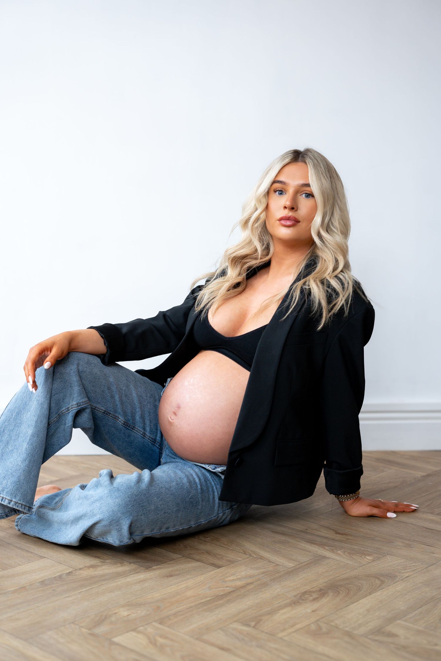 Pregnant woman in black blazer and blue jeans sitting on floor with bare bump in white studio.
