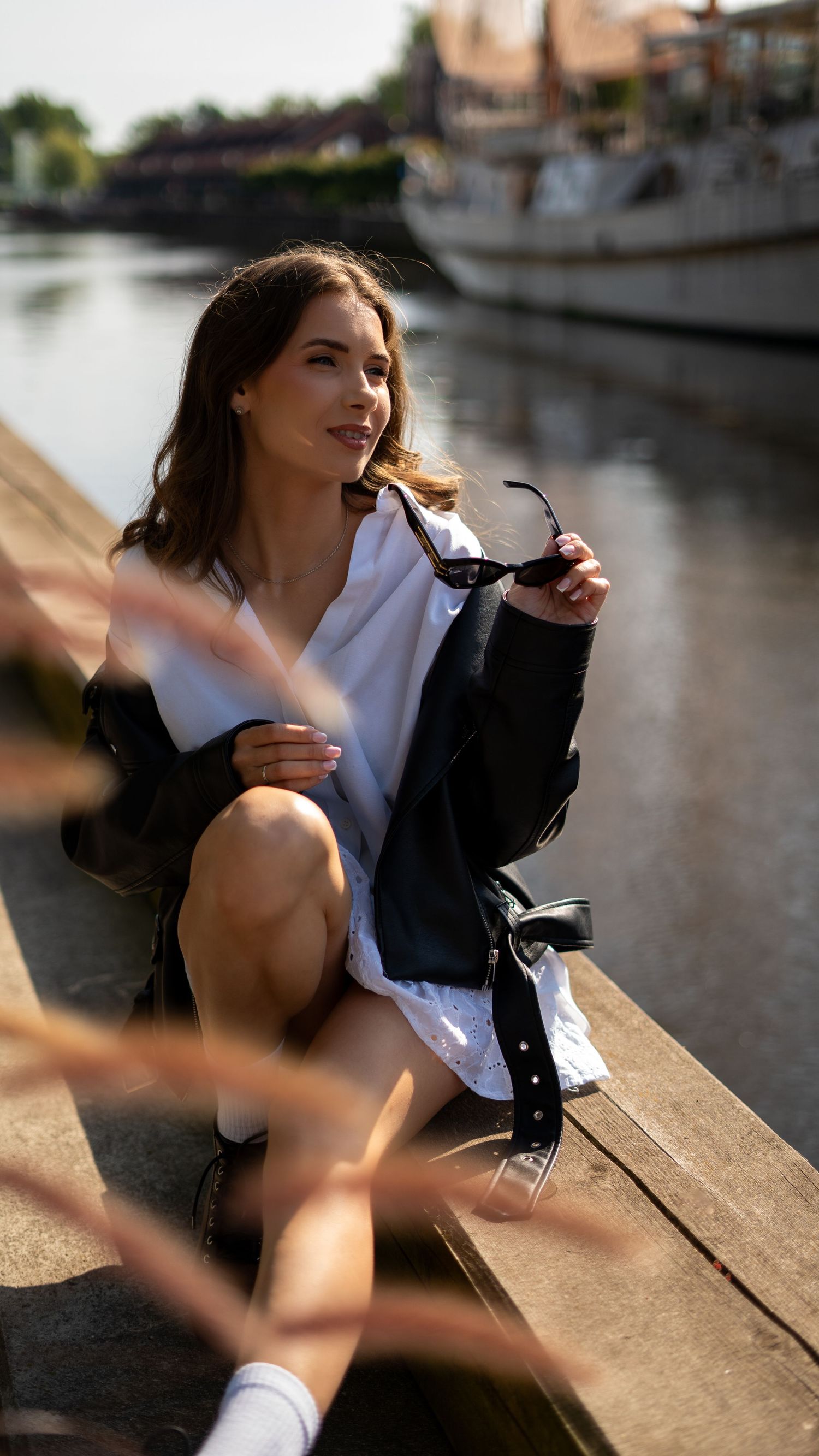 Young woman in black leather jacket and white dress sitting on dock holding sunglasses by the water.