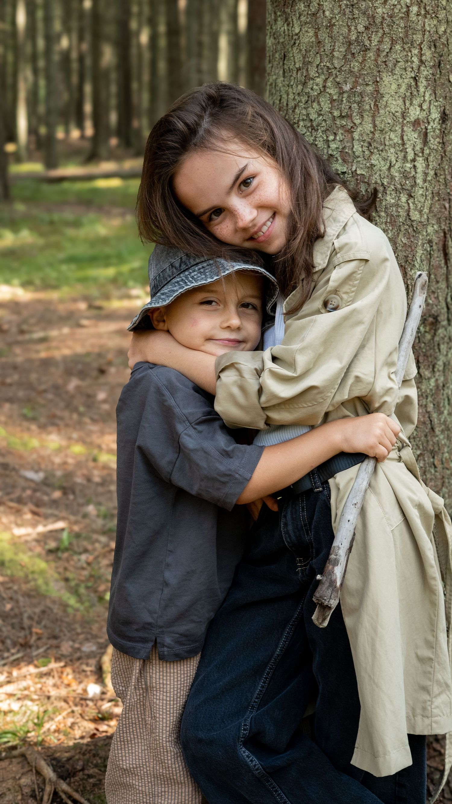 A smiling girl in a trench coat hugs a younger boy wearing a hat against a tree in a forest.
