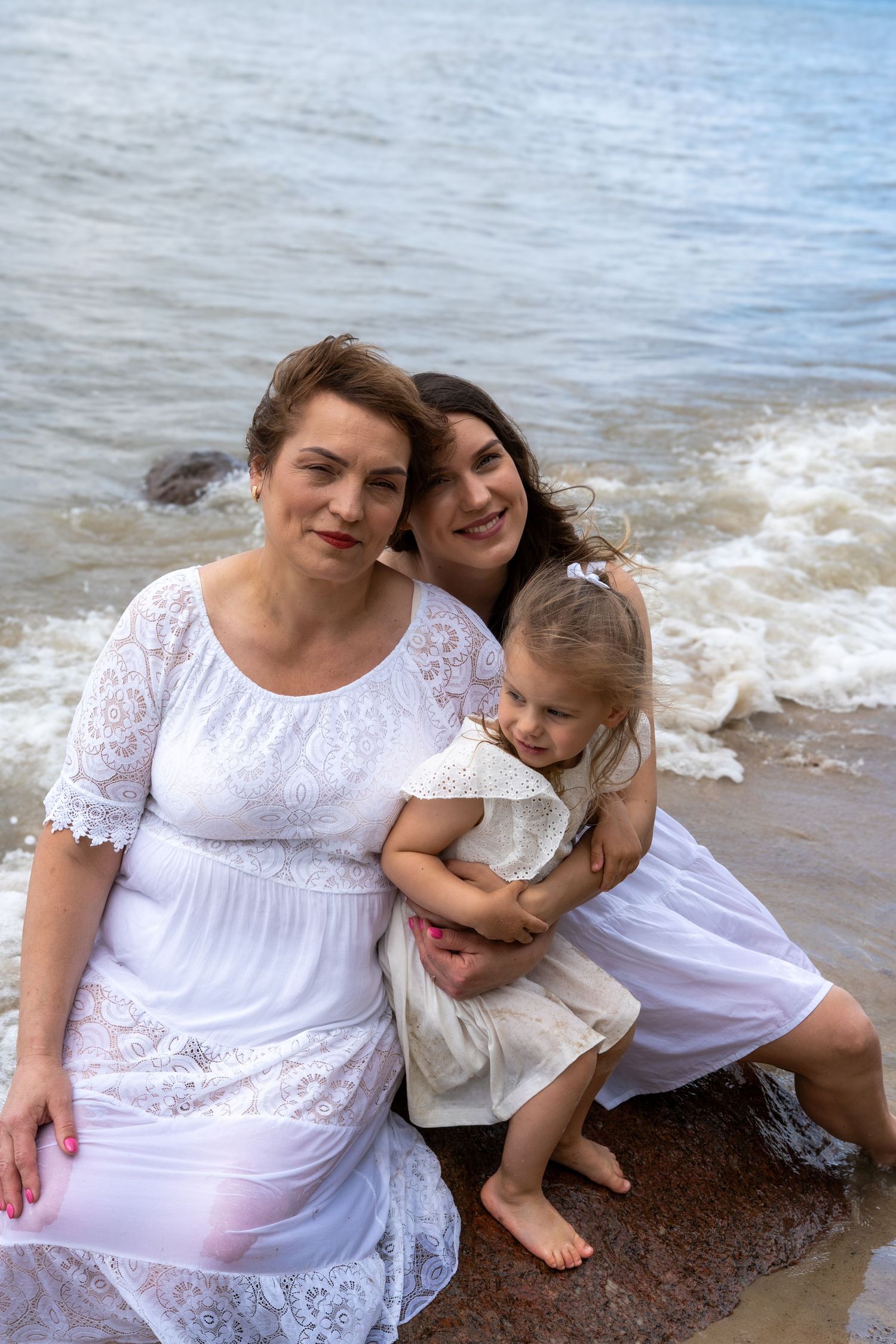 Three generations of women in white dresses pose together on a rocky beach by the ocean.