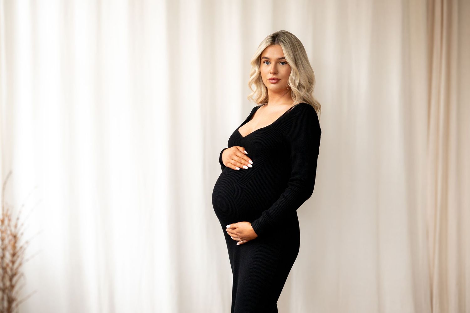 Pregnant woman in fitted black long-sleeve maxi dress cradling baby bump against white curtains.