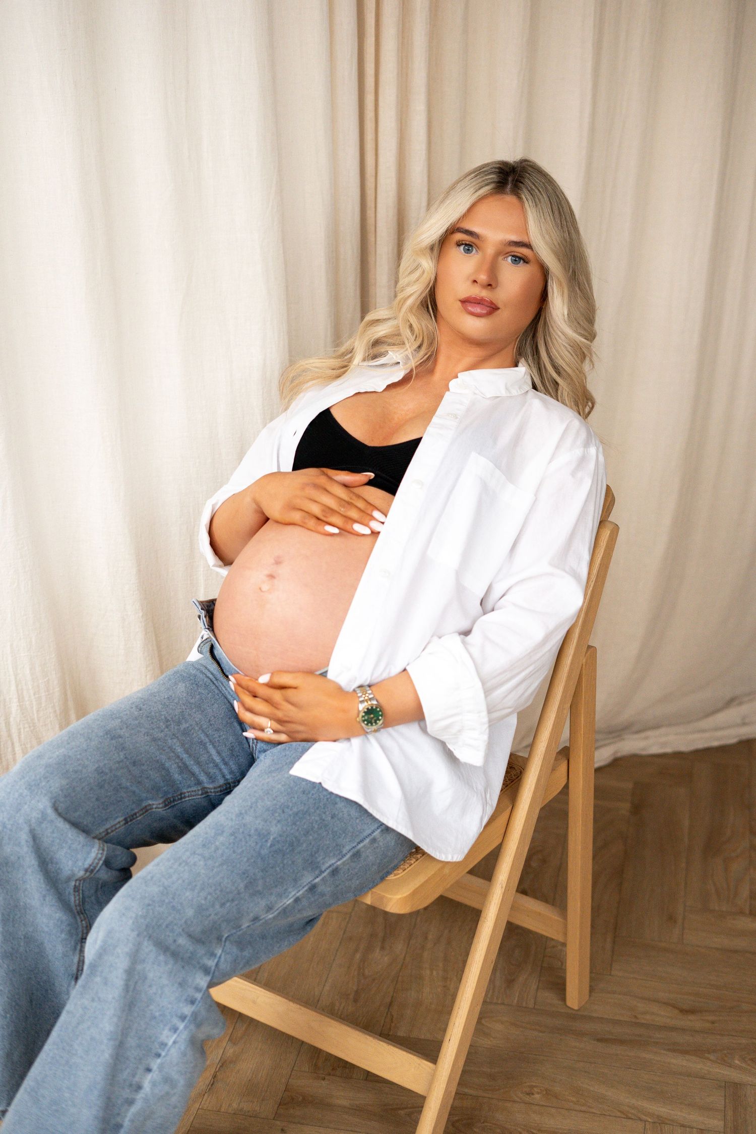 Pregnant blonde woman in white shirt sitting on wooden chair cradling baby bump.