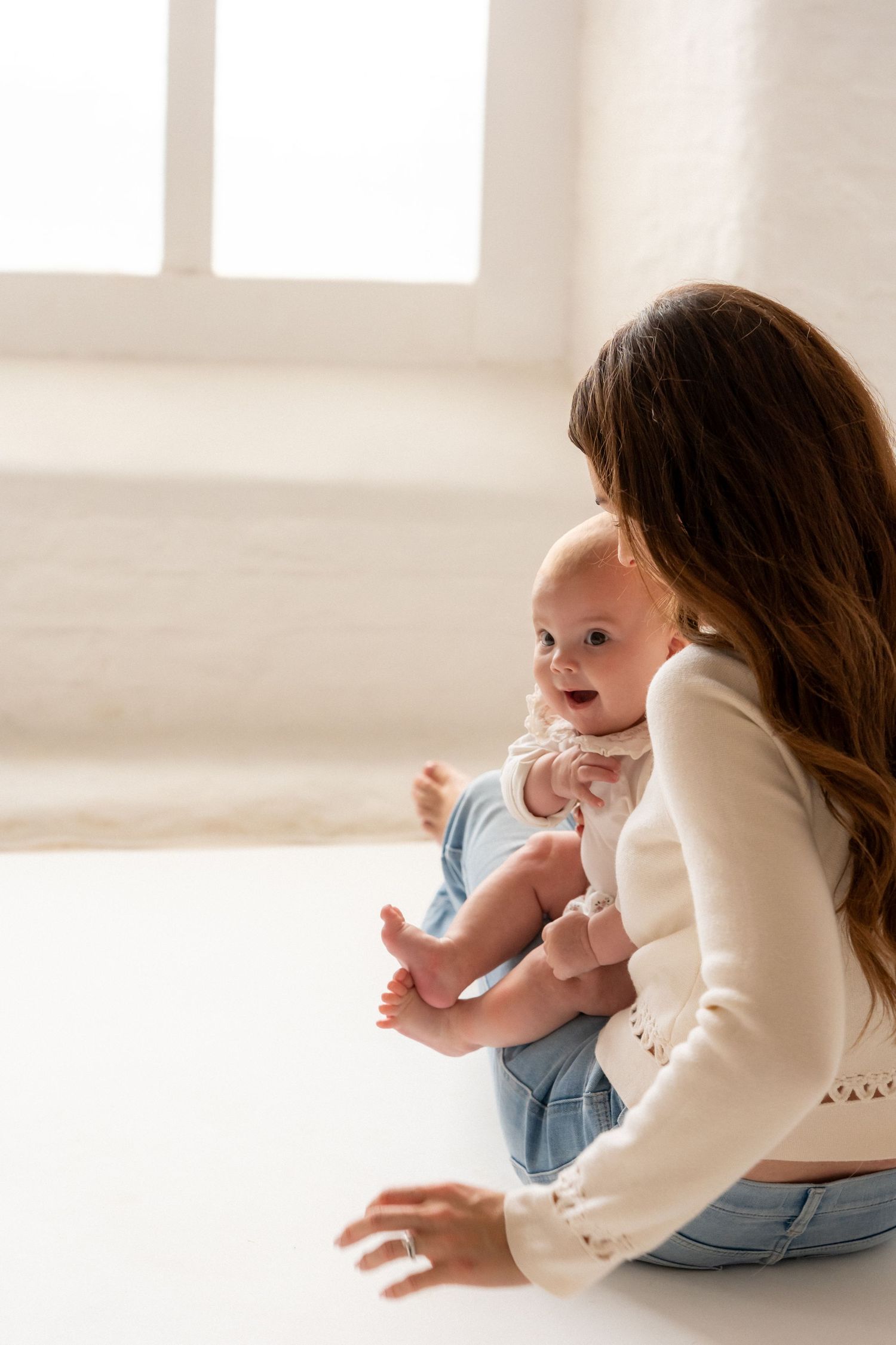 A mother sitting on a white floor holding her smiling baby dressed in blue, with a bright window in the background.