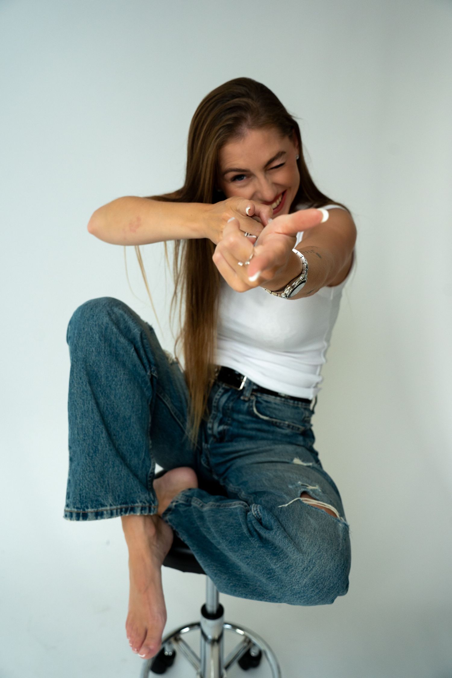 Playful woman in white tank top and ripped jeans points at camera while sitting on chrome stool smiling.