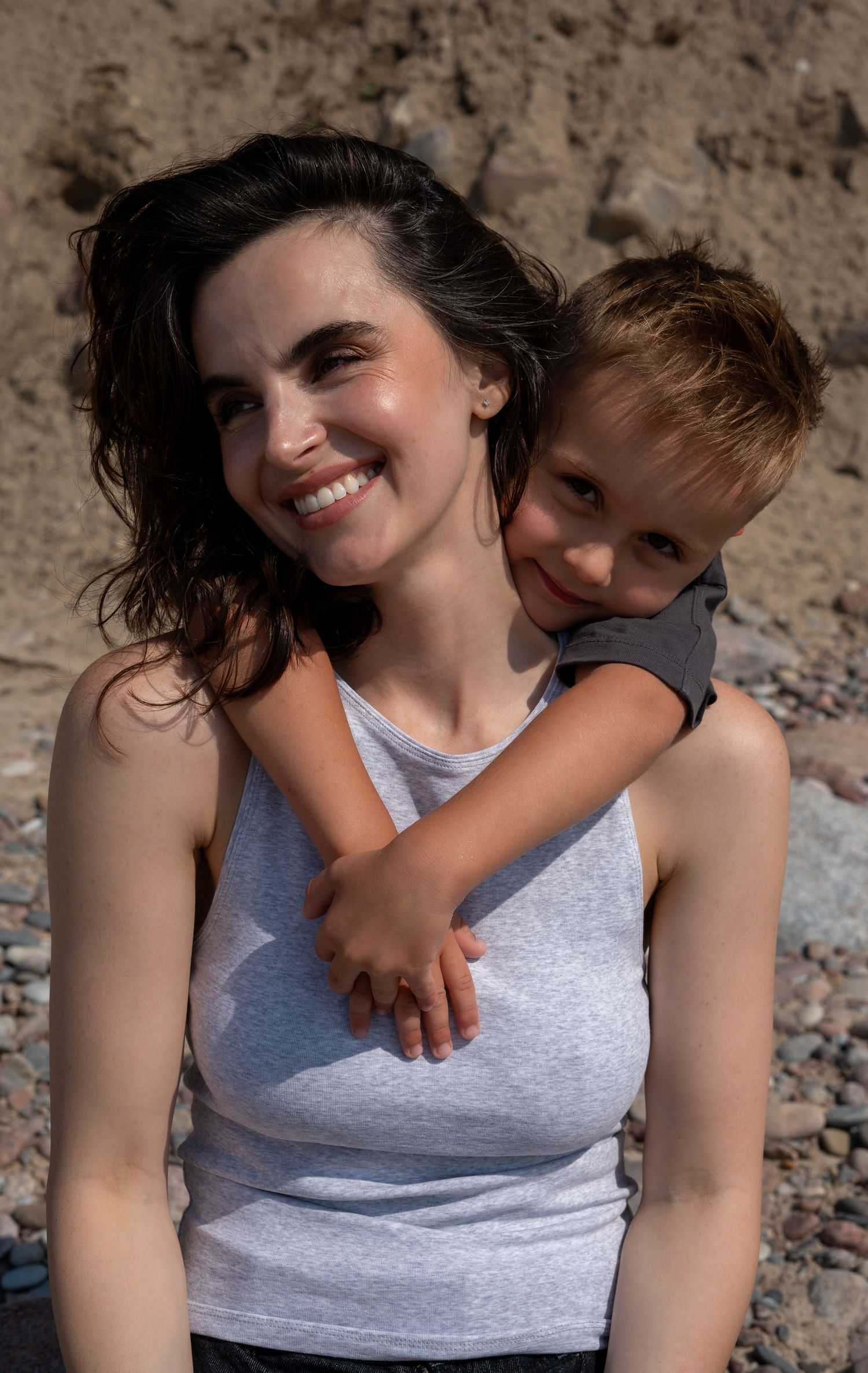 A smiling woman in a white tank top with a young boy hugging her from behind on a rocky beach.