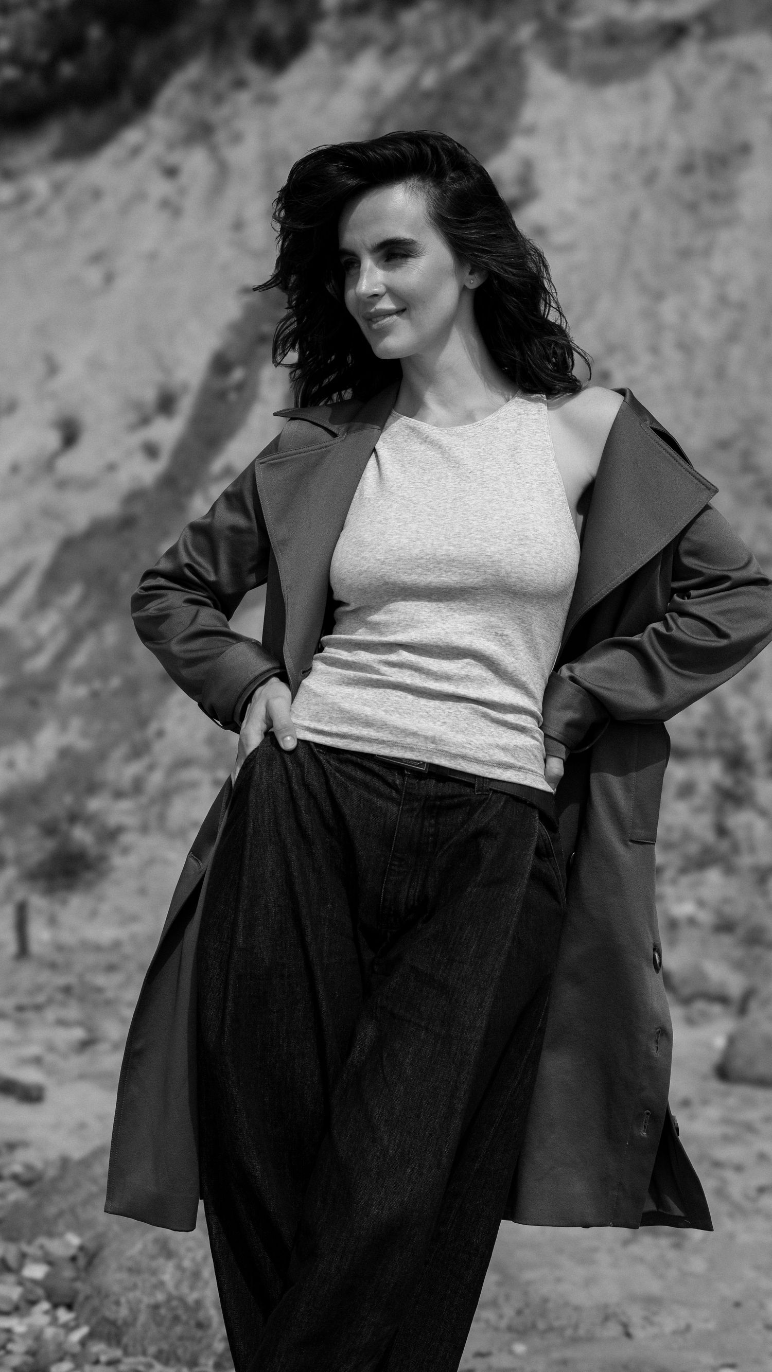Black and white photo of confident woman in long coat and tank top posing outdoors on rocky terrain.