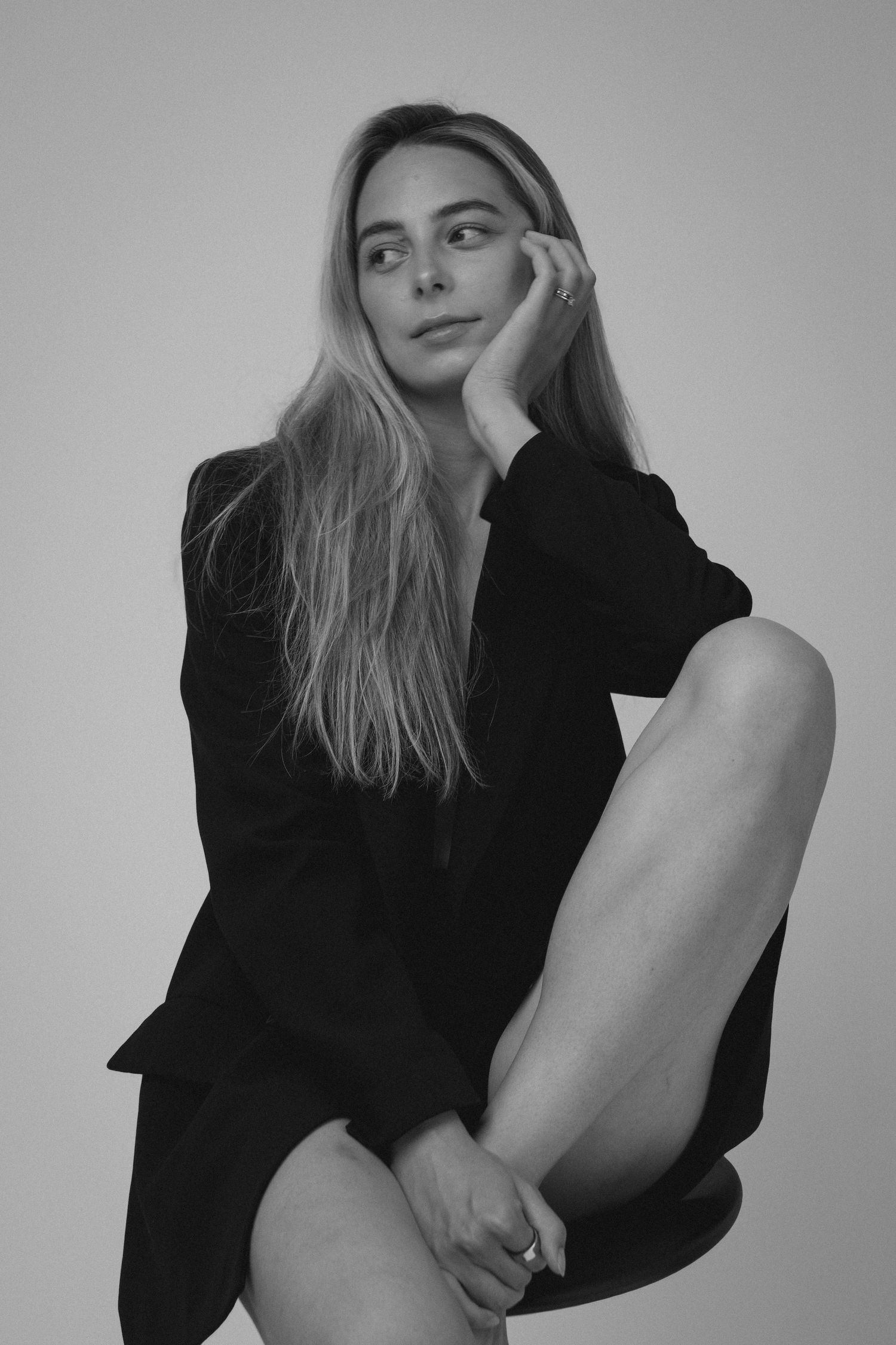 Black and white studio portrait of blonde woman in black blazer sitting and resting hand on face.