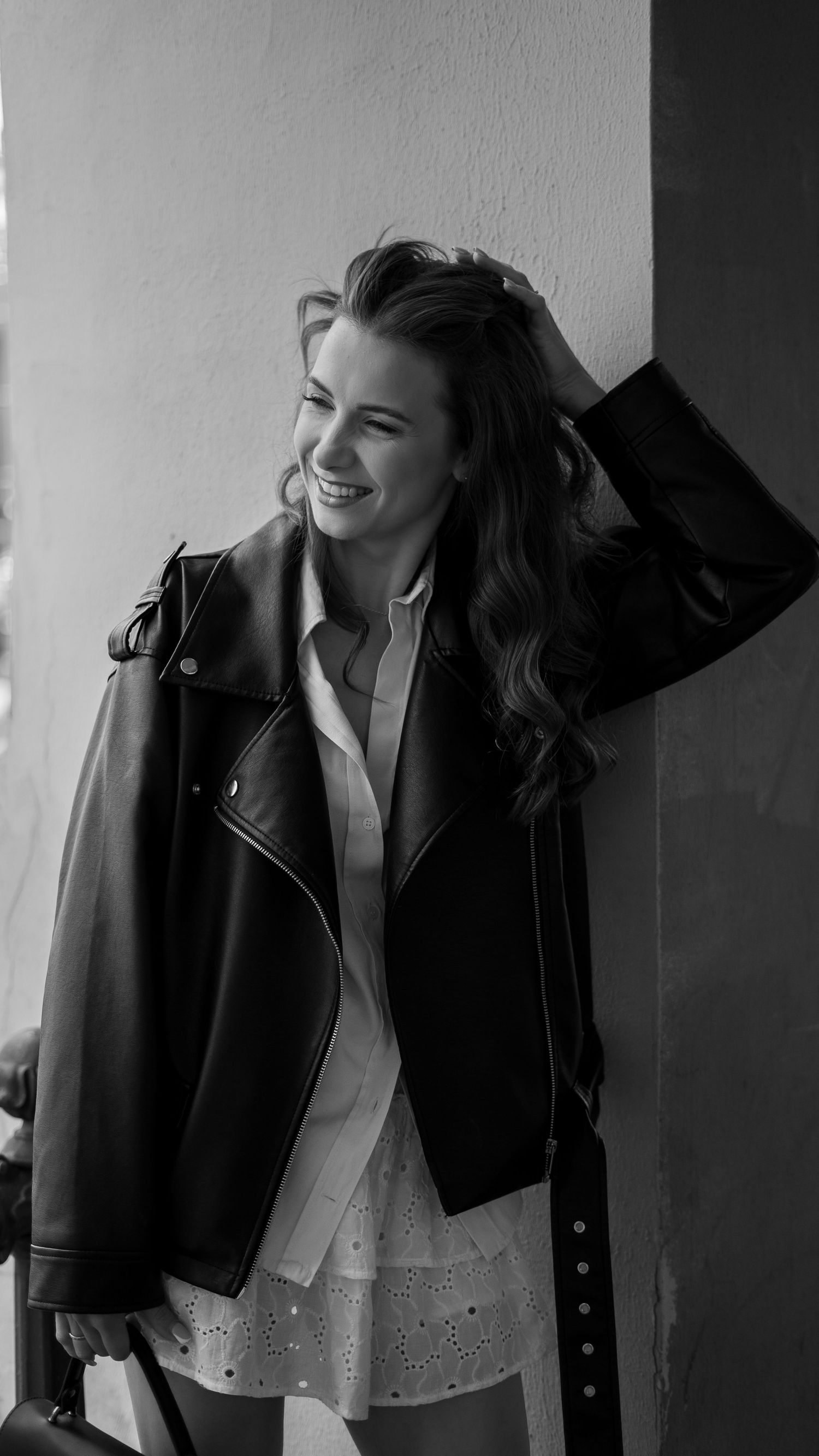 Smiling woman in black leather jacket and white dress leaning against wall in black and white portrait.