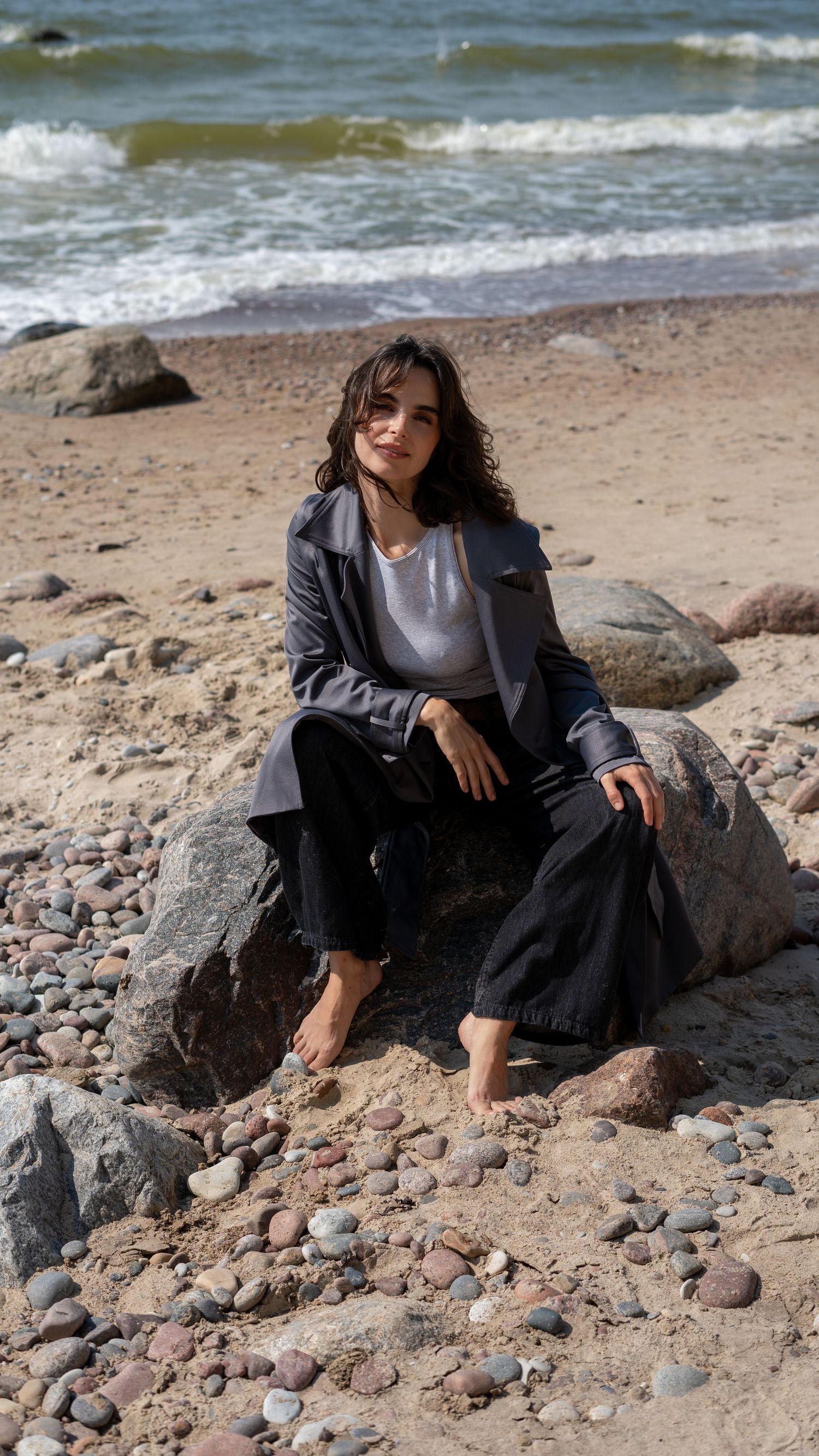 Barefoot woman in dark jacket and pants sitting on rocky beach with waves behind her.