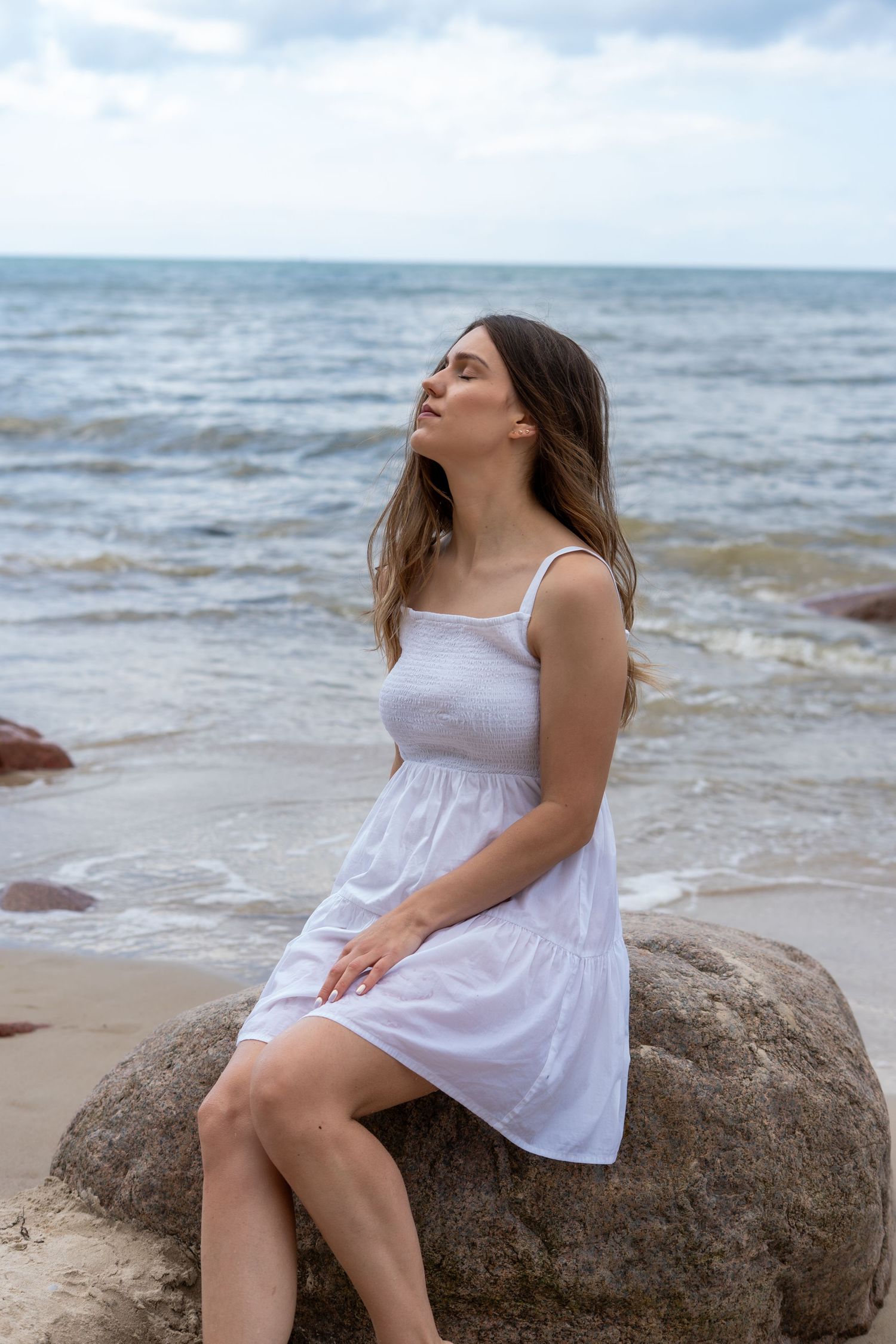 Young woman in white sundress sitting on seaside rock with eyes closed enjoying ocean breeze.