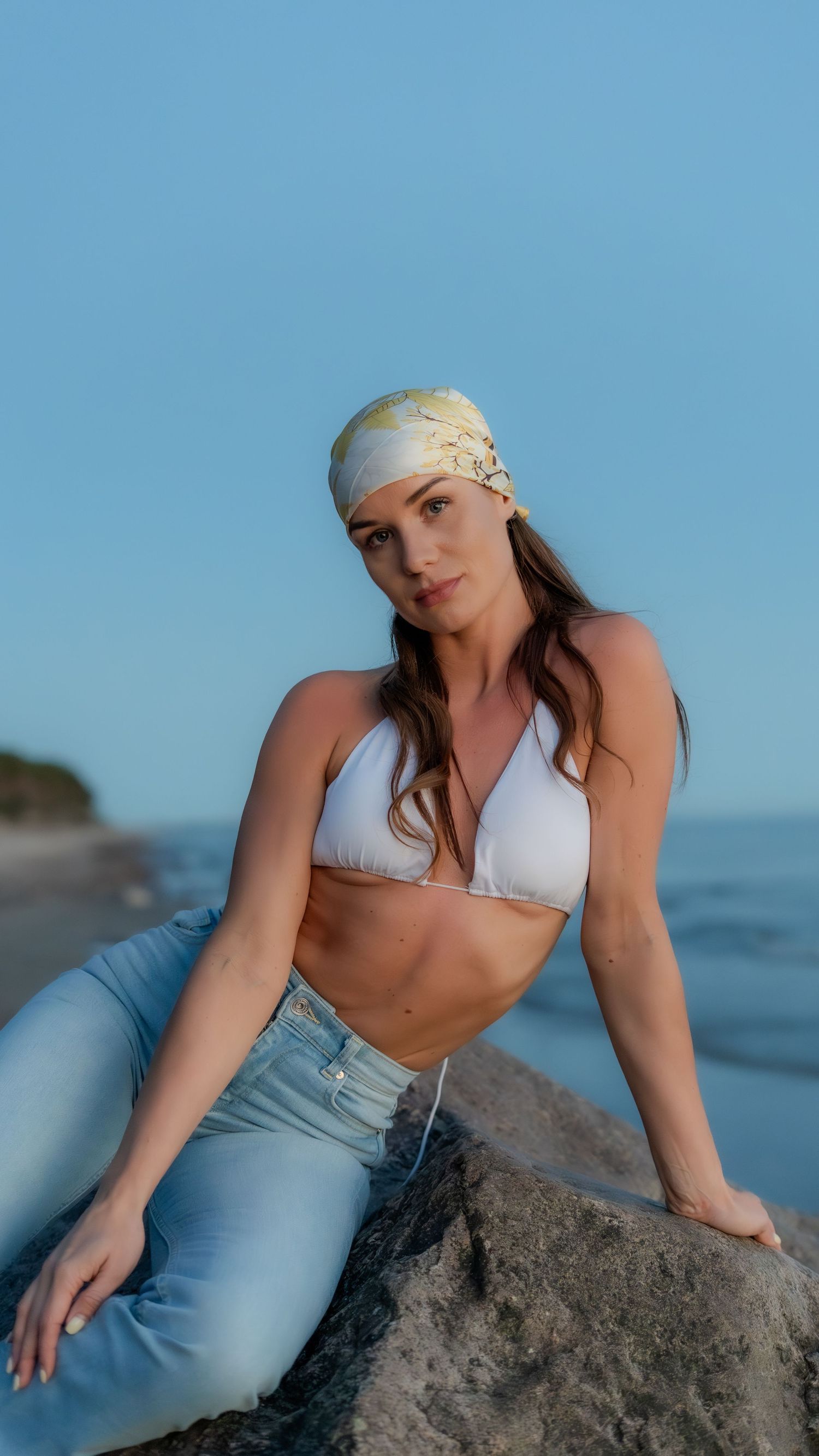 Brunette woman in white halter bikini top and jeans posing on coastal rock.