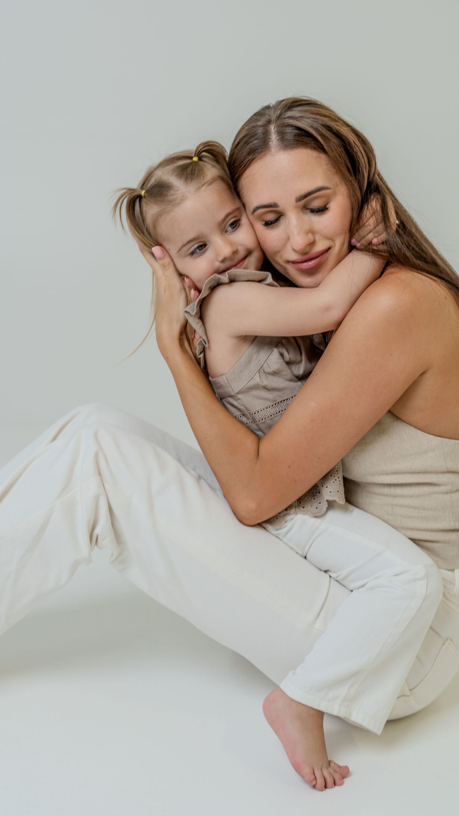 Tender moment of young girl embracing smiling brunette mother in white pants on light gray background.