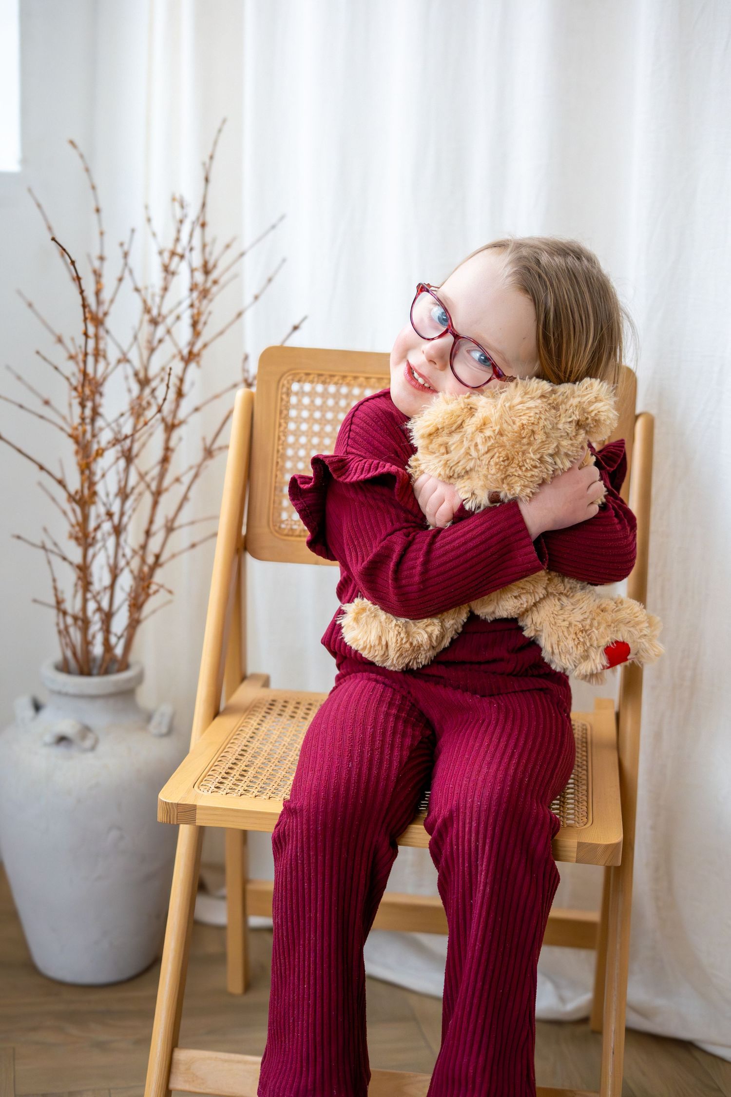 Happy young girl in red glasses and burgundy outfit hugging teddy bear on wooden chair indoors.