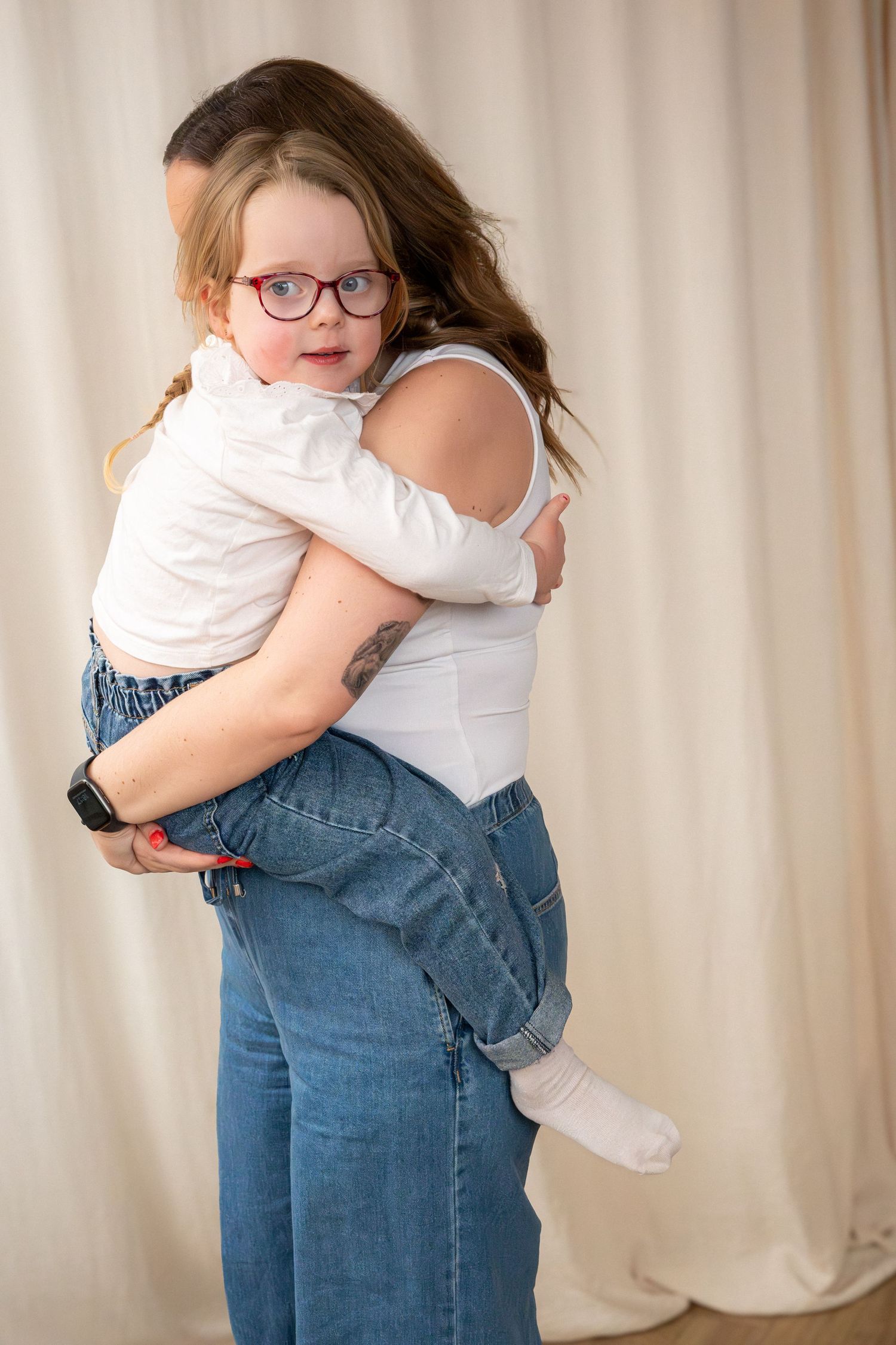 A mother carries her young daughter with glasses in her arms, both smiling warmly.