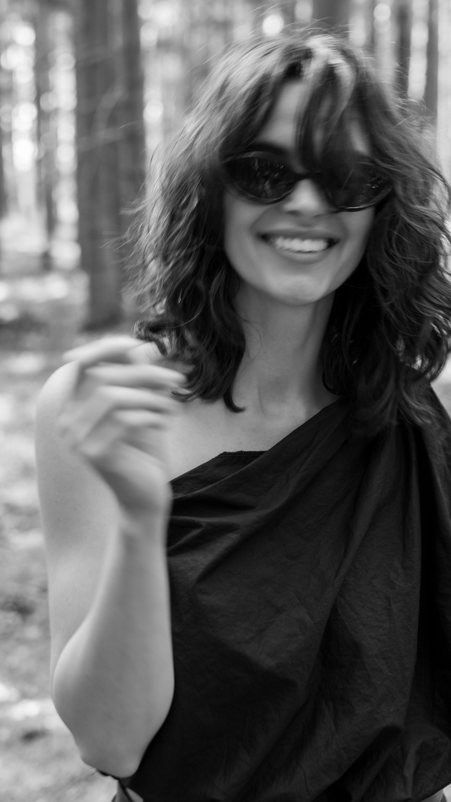 Black and white portrait of smiling woman in one-shoulder top wearing sunglasses in forest setting.
