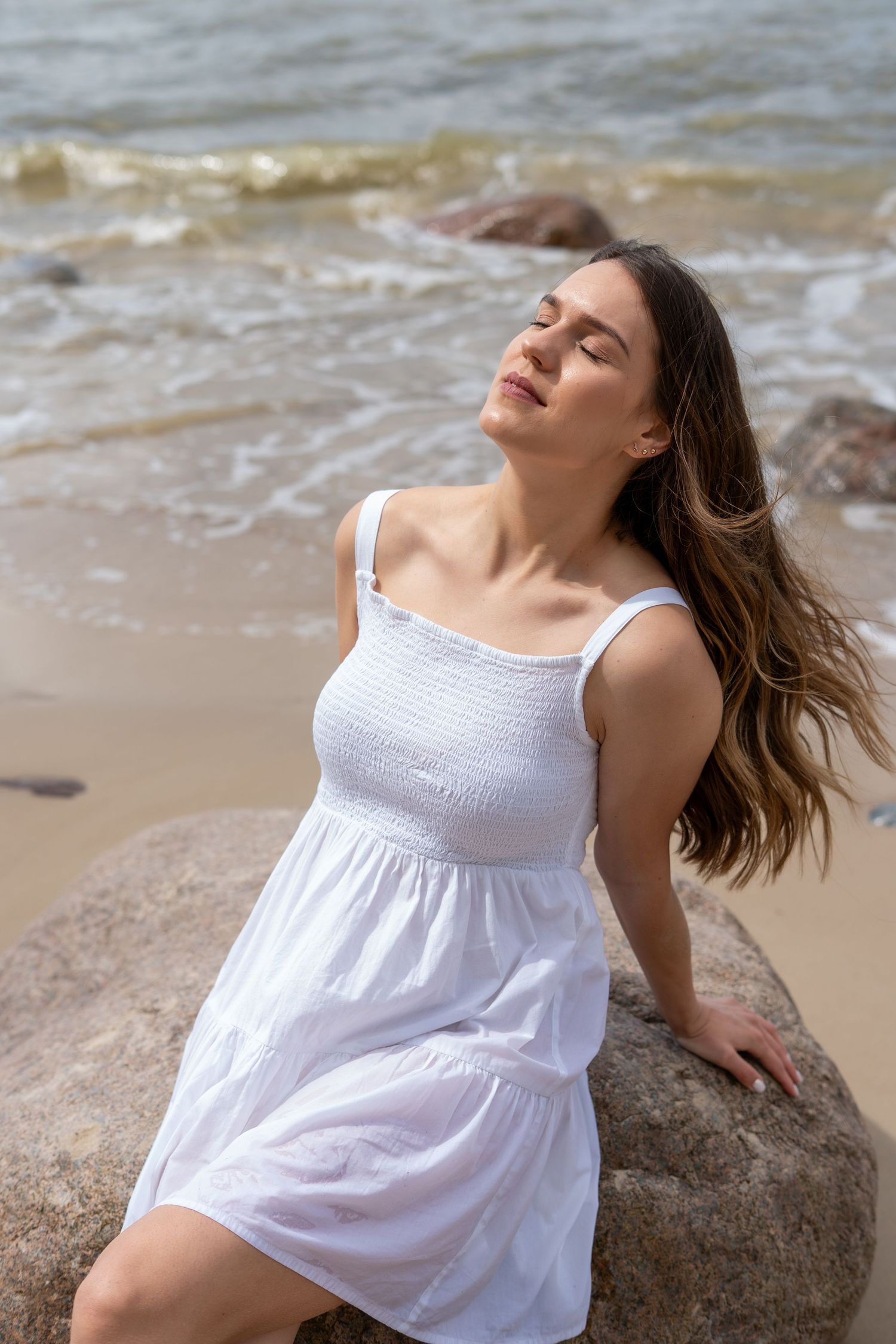 Serene woman in white summer dress leaning on boulder at beach with eyes closed in sunlight.