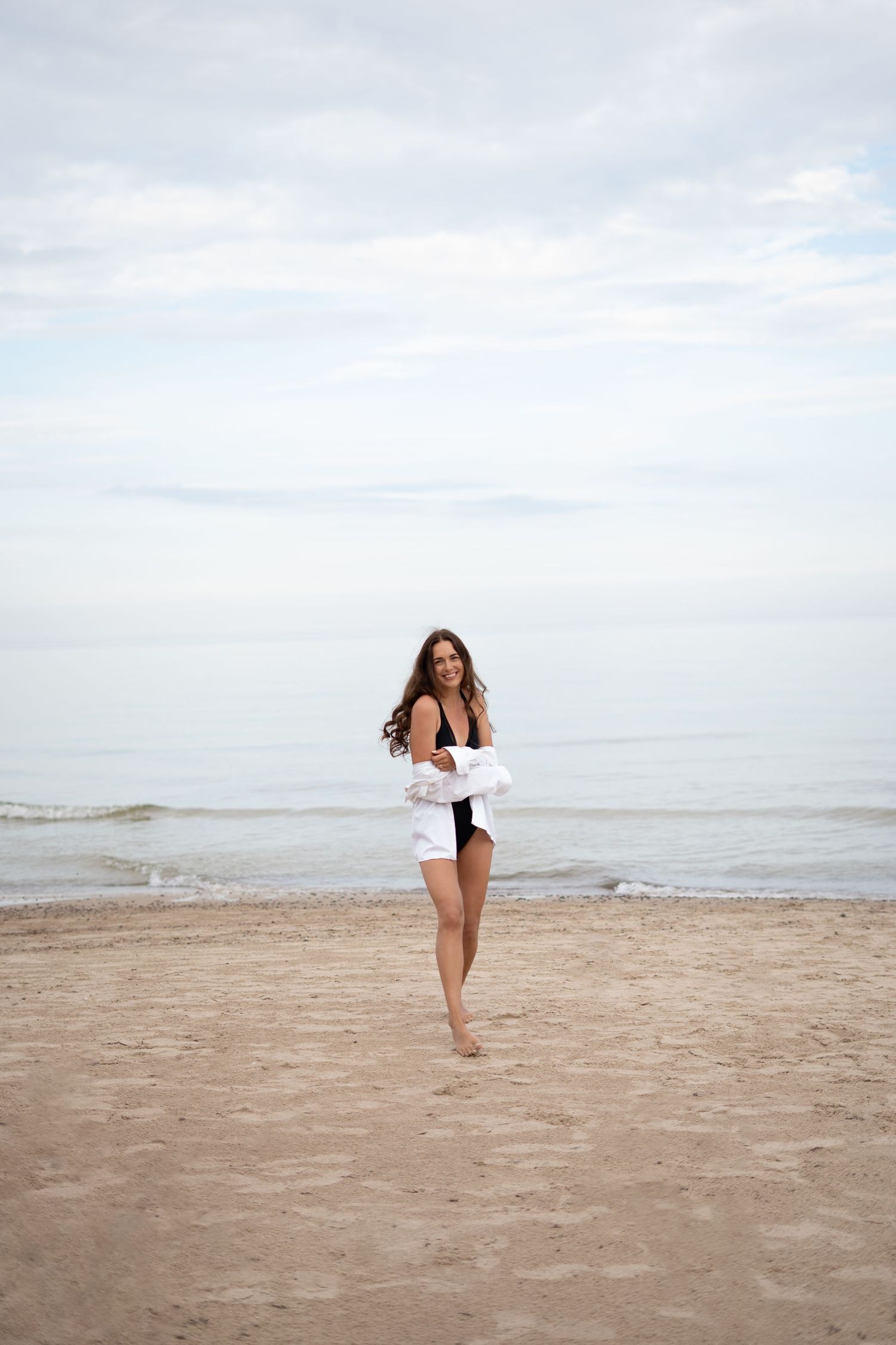 Brunette woman in black swimsuit and white shirt walking barefoot on sandy beach by the sea.