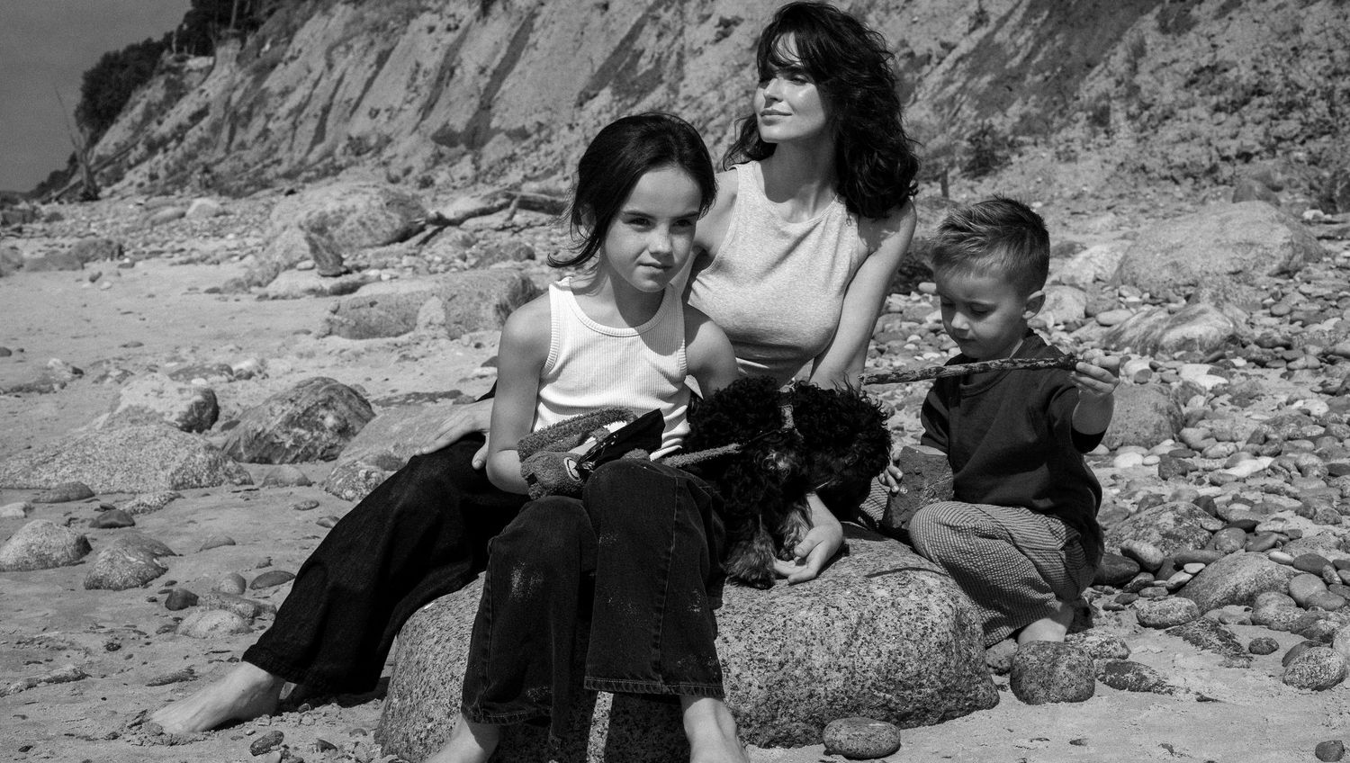 Black and white photo of a mother and two children sitting on a rocky beach with cliffs in the background.