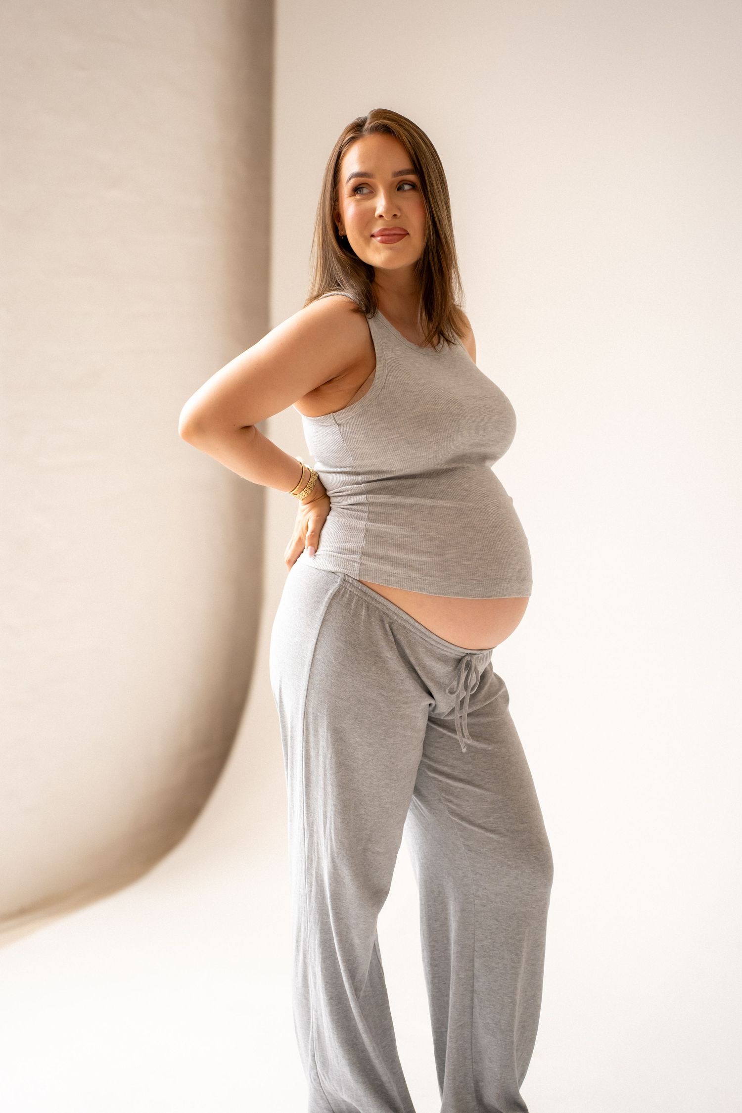 Pregnant woman in gray sleeveless top and loose pants posing with hand on hip, showcasing baby bump.