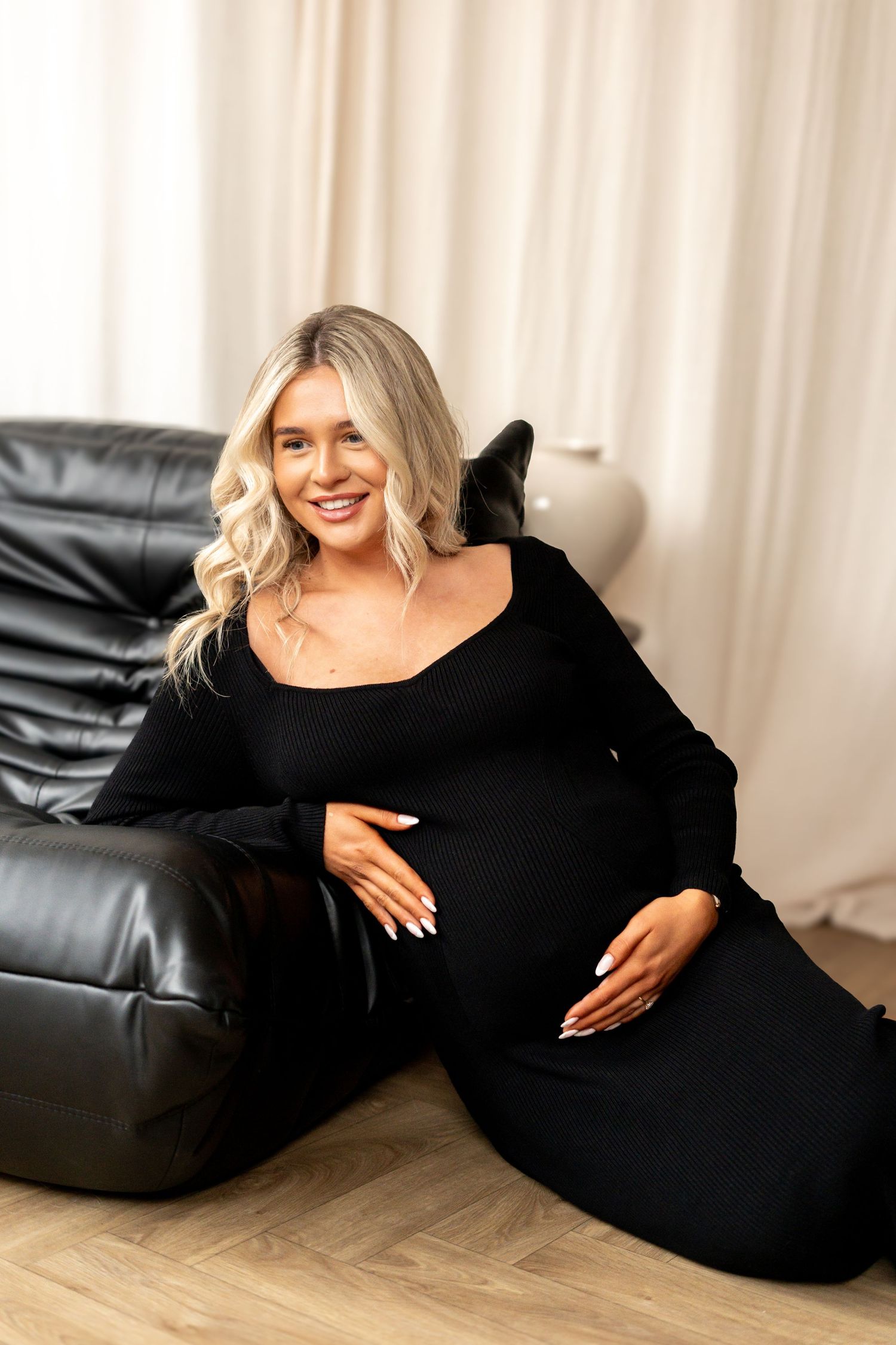 Smiling pregnant woman in black dress sitting on floor leaning against black leather sofa indoors.